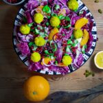 Eine Platte mit Radicchio-Orangen-Salat, der mit in Pistazienmehl panierten Ricottabällchen belegt ist. Eine Orange, eine angeschnittene Zitrone und ein Schüsselchen mit Granatapfelkernen sind um den Teller platziert.