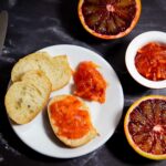 Sizilianische Blutorangenmarmelade: Teller mit Brotscheiben, eine mit Konfitüre bestrichen. Daneben aufgeschnittene Blutorangen und ein Töpfchen mit Marmelade.