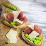 Ein Holzbrett, auf dem zwei mit Crostini liegen, die mit Erbsencreme bestrichen und mit Bresaolastreifen und Parmesanhobeln belegt sind.