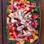 Eine Salatplatte mit Rucola, Melonenstücken, Garnelen und Bresaolastreifen, von oben aufgenommen.