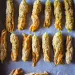 Mehrere Reihen von gefüllten, panierten Zucchiniblüten auf einem mit Backpapier ausgelegten Backblech.