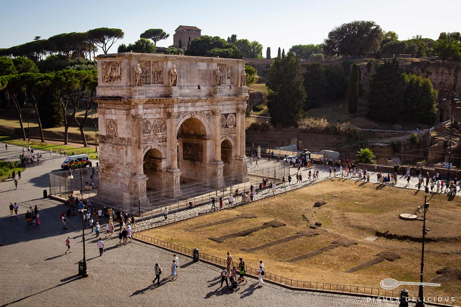 Der Konstantinsbogen in Rom. Die gut erhaltene Sehenswürdigkeit mit drei Durchgängen ist mit vielen Statuen und steineren Reliefs verziert. Im Hintergrund ist das Forum Romanum zu sehen, rund um den Bogen spazieren Touristen.