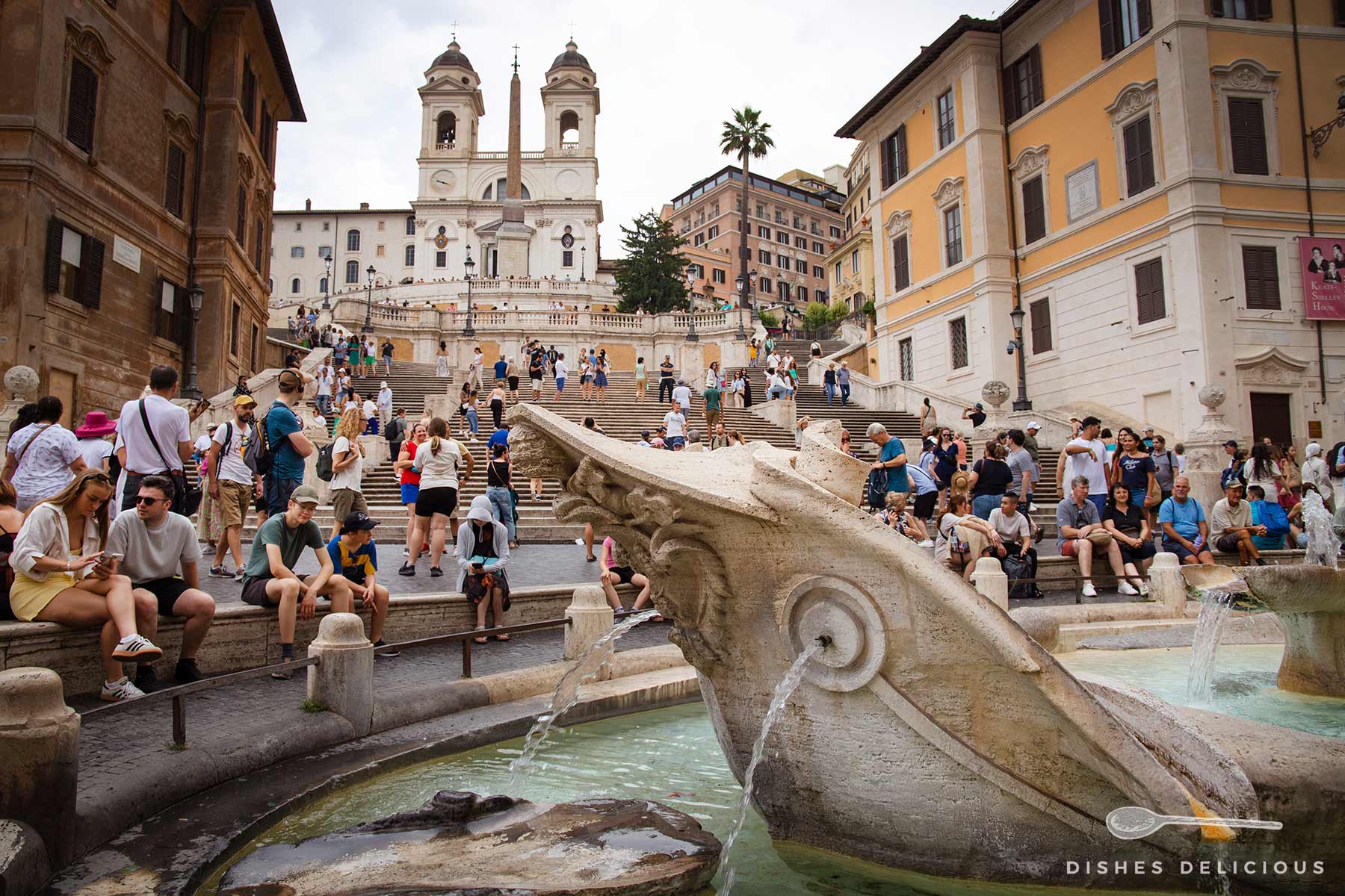 Zahlreiche Touristen rund um die Fontana della Barcaccia an der Spanischen Treppe. Im Hintergrund sind die berühmten Zwillingstürme der Trinità dei Monti Kirche sichtbar, während Menschen die steilen Stufen hinauf und hinabgehen.