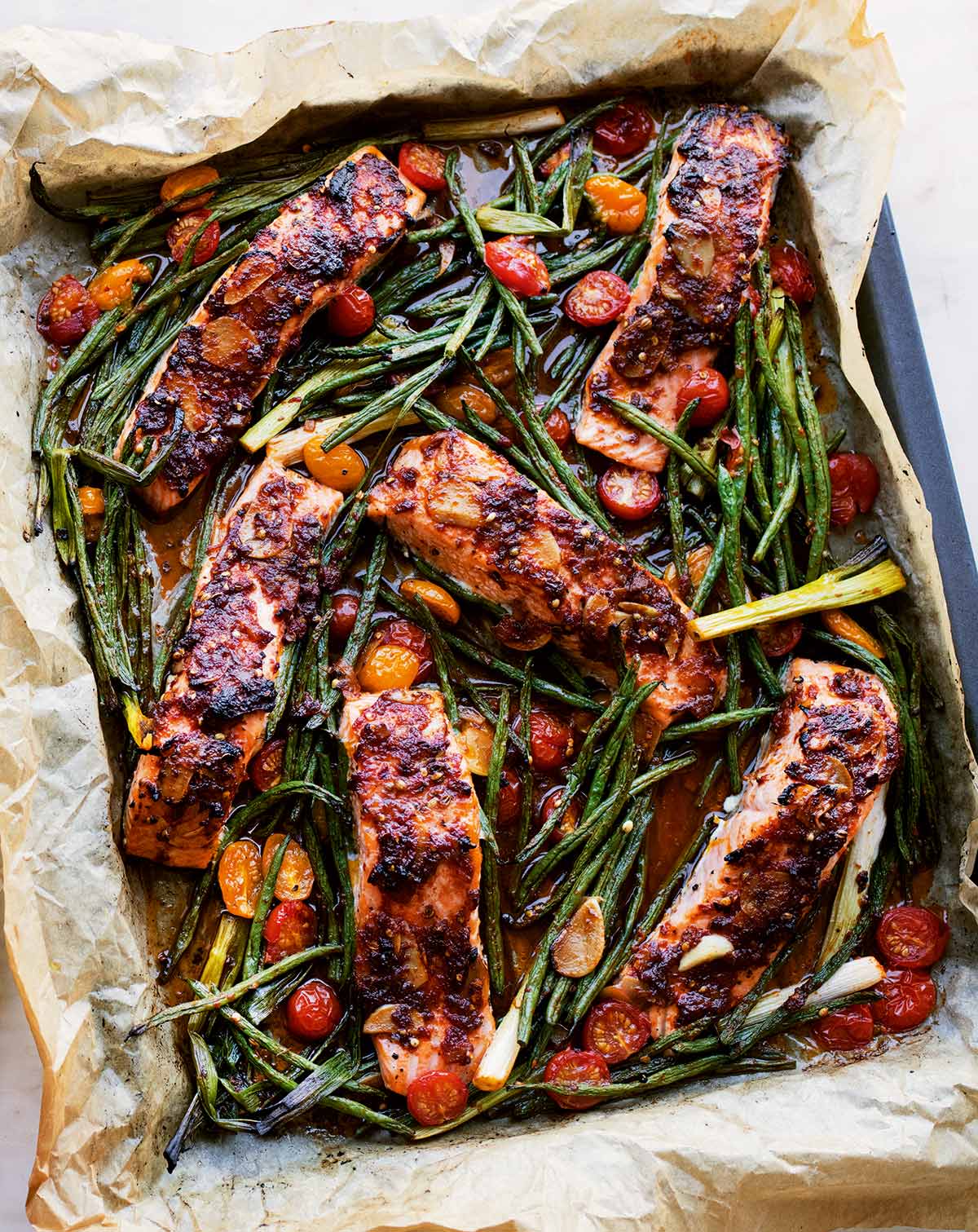 Ein Backblech mit sechs knusprig gegarten Lachfilets mit Bohnen und Kirschtomaten.