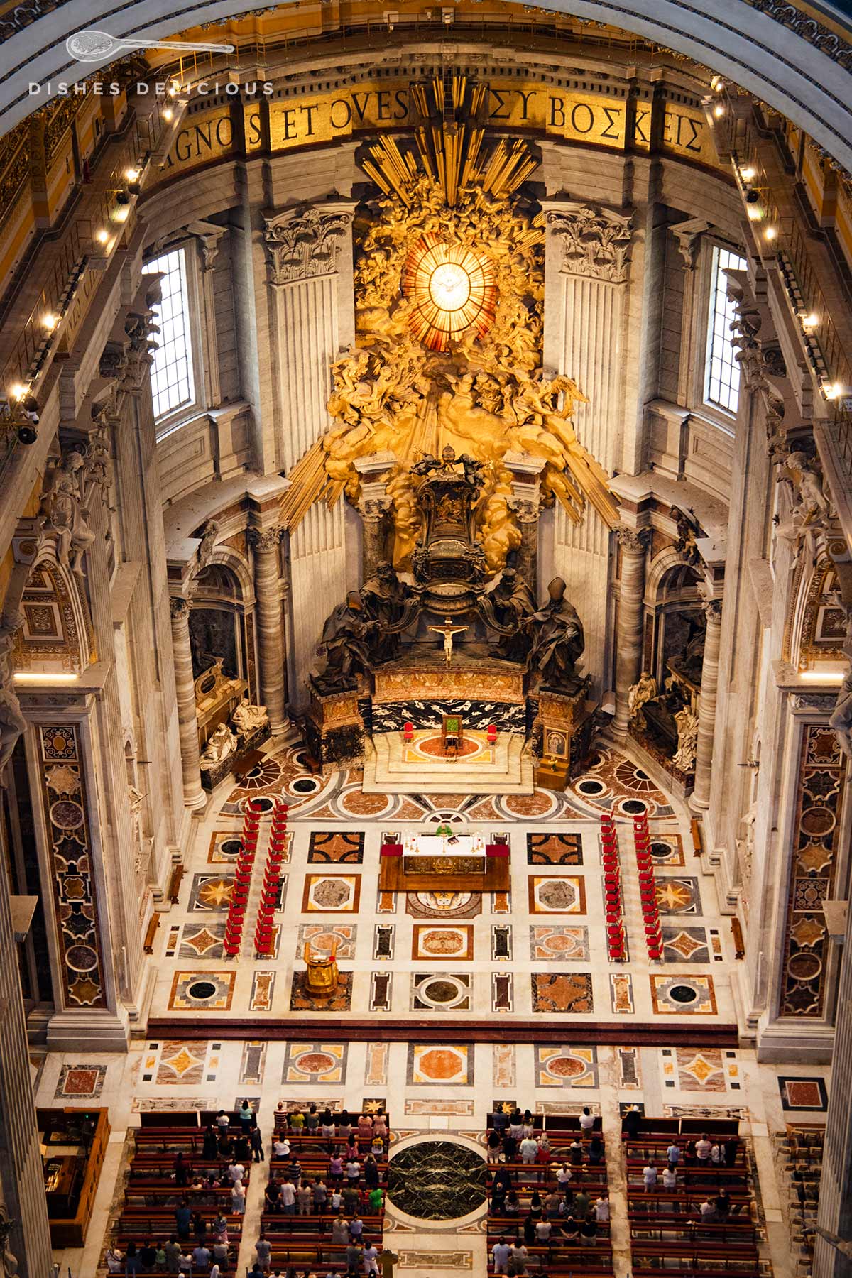 Der spektakuläre Altar Cathedra Petri im Petersdom von der Kuppel aus gesehen, davor der mit bunten Mosaiken gestaltete Boden und Kirchenbänke.