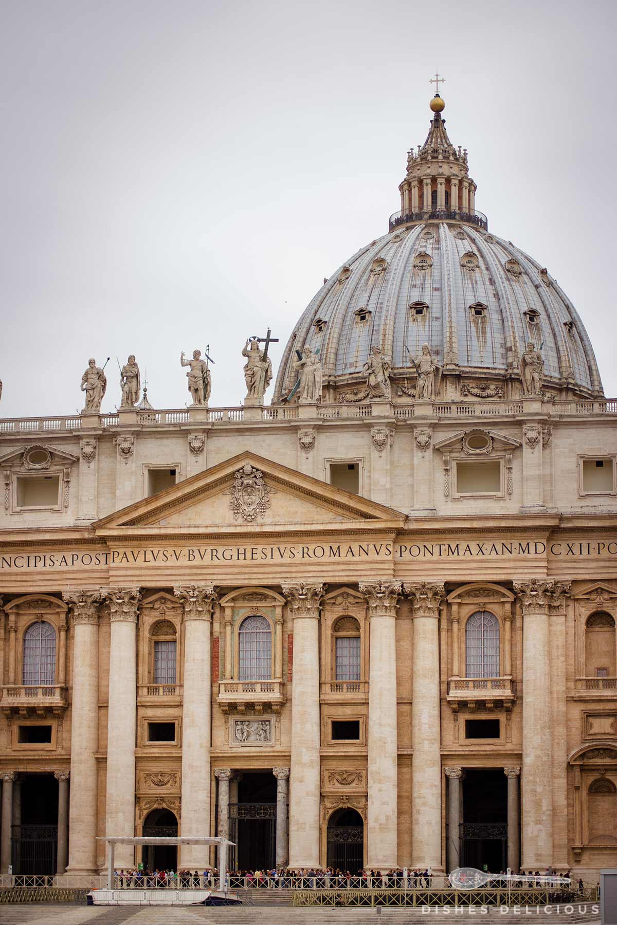 Das Bild zeigt die Fassade der Petersbasilika in Rom mit einer großen Kuppel und mehreren Statuen auf dem Dach. Darunter befinden sich beeindruckende Säulen und ein Schriftzug. Im Vordergrund ist eine Warteschlange von Menschen zu sehen.