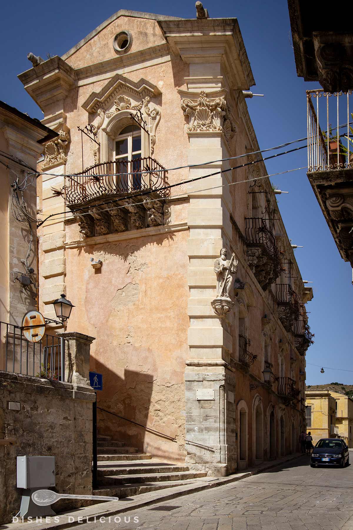 Seitenansicht des Palazzo Cosentini: ein barockverziertes Fenster mit Balkon im Obergeschoss. Eine Statue und Verzierungen sind an der Ecke des Gebäudes angebracht.