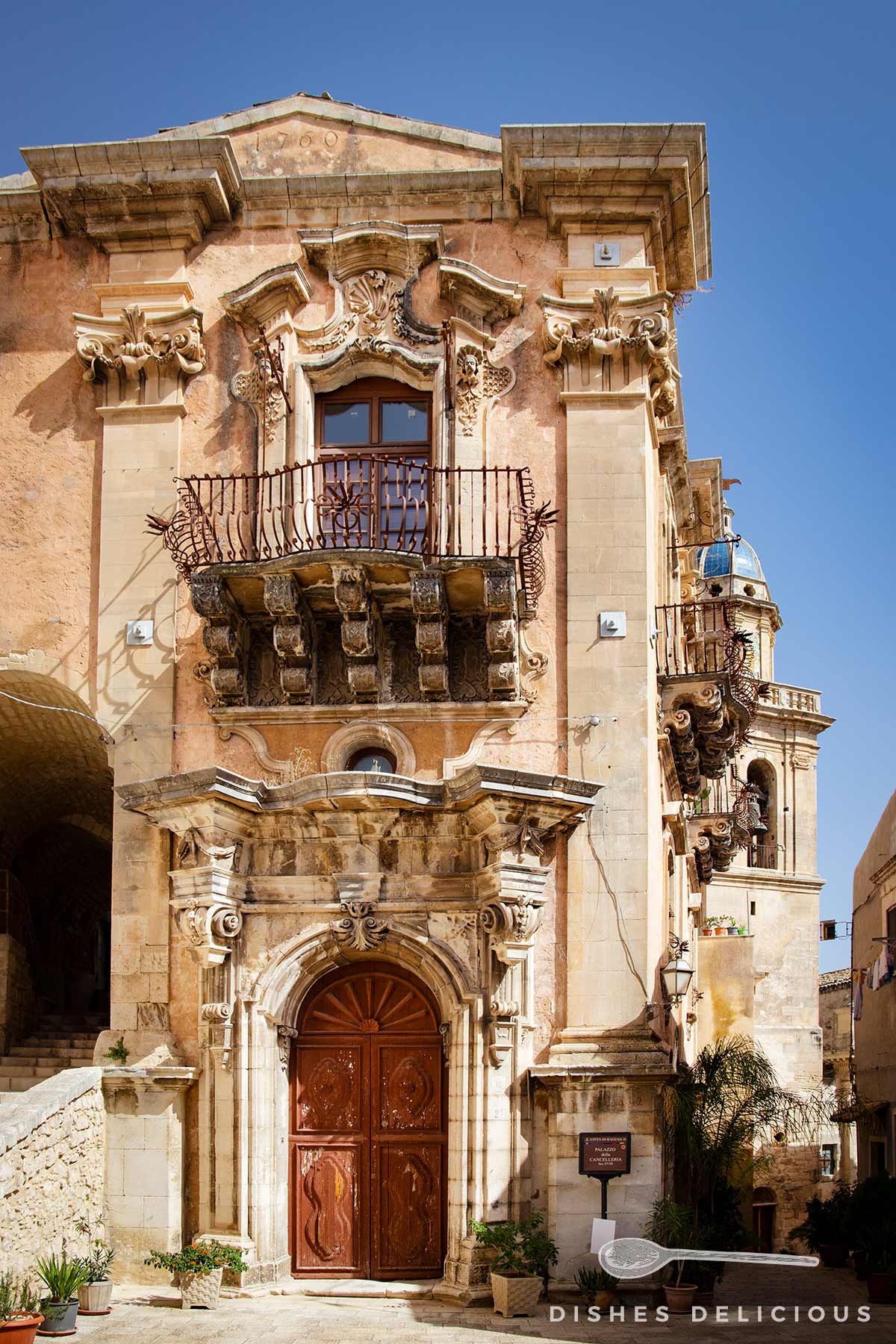 Palazzo della Cancelleria in Ragusa: alter Barockpalast mit zwei Etagen, einem prachtvoll verzierten Eingangsportal und einem reich verzierten Fenster mit Balkon in der zweiten Ebene.