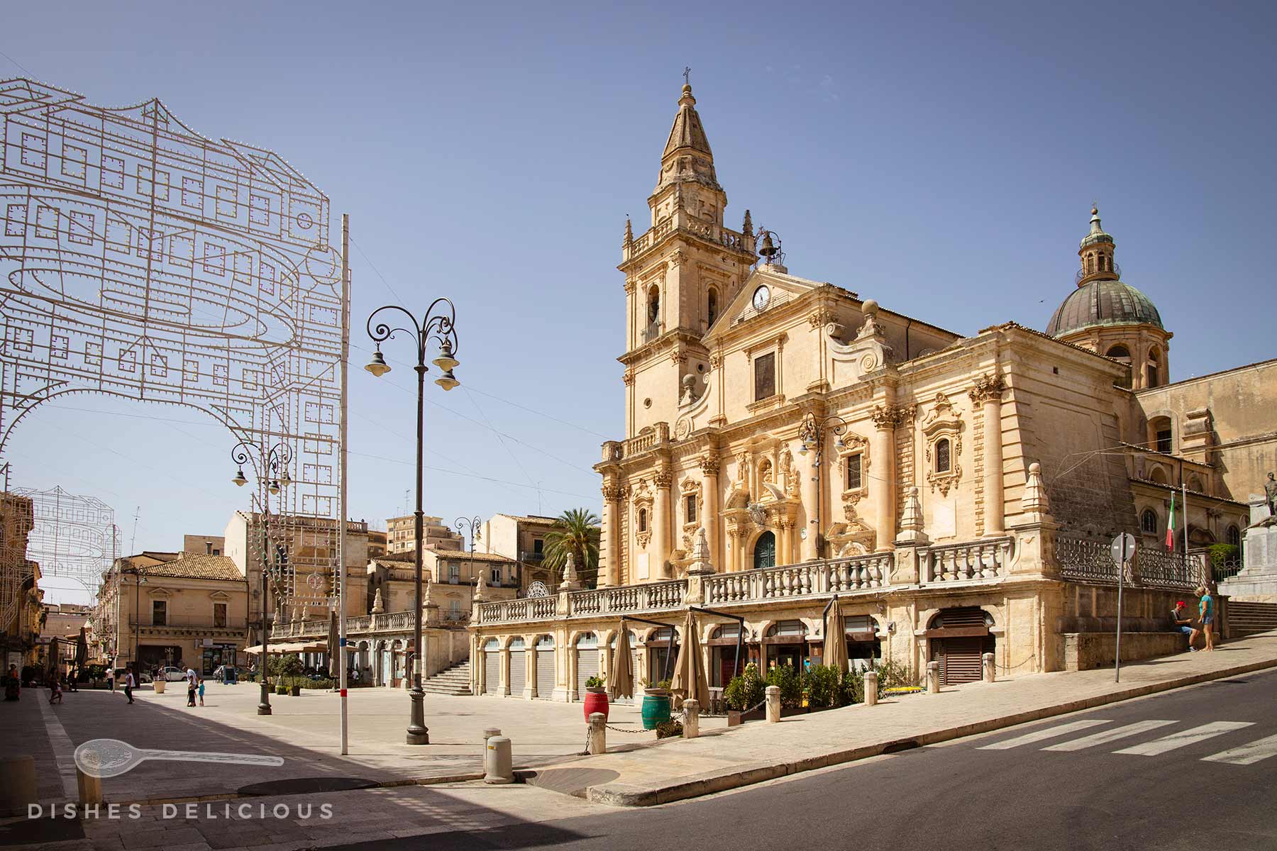 Die barocke Kathedrale San Giovanni am sonnigen Hauptplatz von Ragusa, Sizilien. Im Vordergrund laden Straßencafés zum Verweilen ein, während dekorative Lichterbögen auf ein Fest hindeuten.