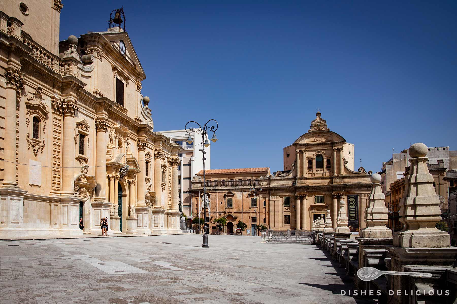 Platz vor der barocken Kathedrale in Ragusa Superiore und die Badia-Kirche. Die Gebäude sind hellbeigefarben und haben eine klassische Architektur mit Barockverzierungen.