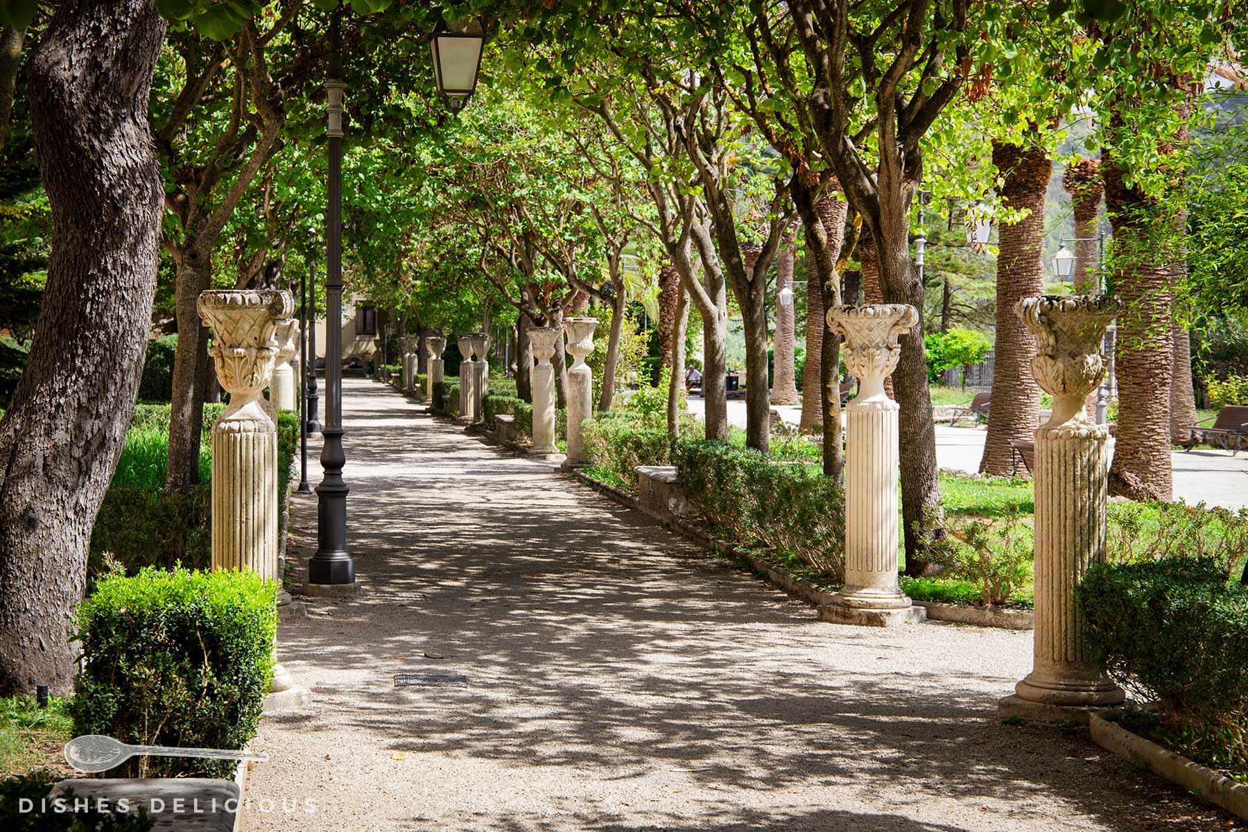 Giradino Ibleo in Ragusa: ein Weg im Park, der von Bäumen und Sträuchern gesäumt ist sowie von mittelhohen Säulen im antiken Stil mit Vasen.