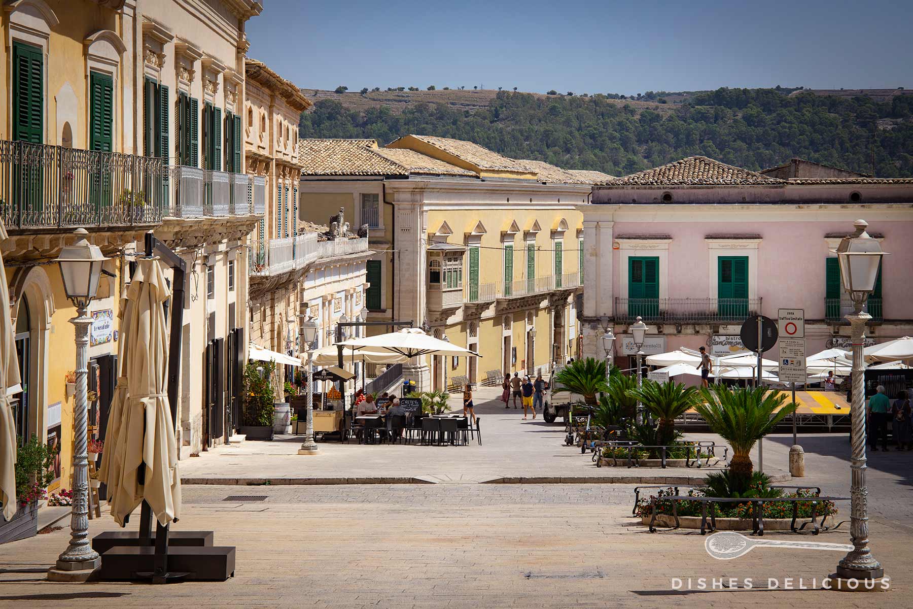 Domplatz und Corso in Ibla mit Palazzi, vor denen Sonnenschirme, Tische und Stühle von Cafés stehen.