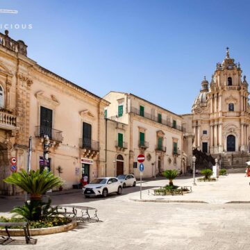 Domplatz von Ragusa Ibla. Im Vordergrund einige Bänke und eine kleine Palme, umgeben von Gebäuden im Barockstil. Im Hintergrund steht die Kathedrale San Giorgio, die den Platz dominiert. Der blaue Himmel und das helle Sonnenlicht unterstreichen die Schönheit des Ortes.