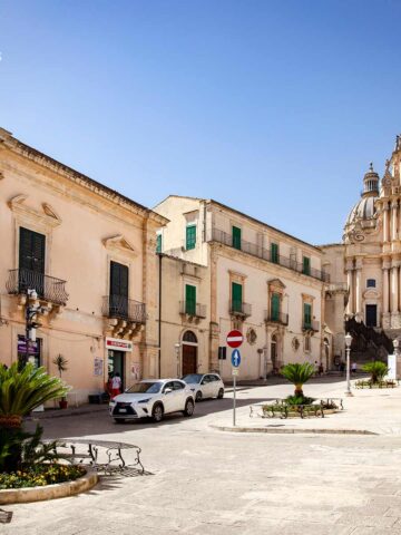 Domplatz von Ragusa Ibla. Im Vordergrund einige Bänke und eine kleine Palme, umgeben von Gebäuden im Barockstil. Im Hintergrund steht die Kathedrale San Giorgio, die den Platz dominiert. Der blaue Himmel und das helle Sonnenlicht unterstreichen die Schönheit des Ortes.
