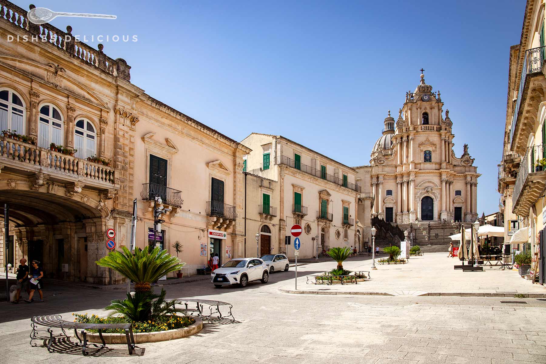 Domplatz von Ragusa Ibla. Im Vordergrund einige Bänke und eine kleine Palme, umgeben von Gebäuden im Barockstil. Im Hintergrund steht die Kathedrale San Giorgio, die den Platz dominiert. Der blaue Himmel und das helle Sonnenlicht unterstreichen die Schönheit des Ortes.