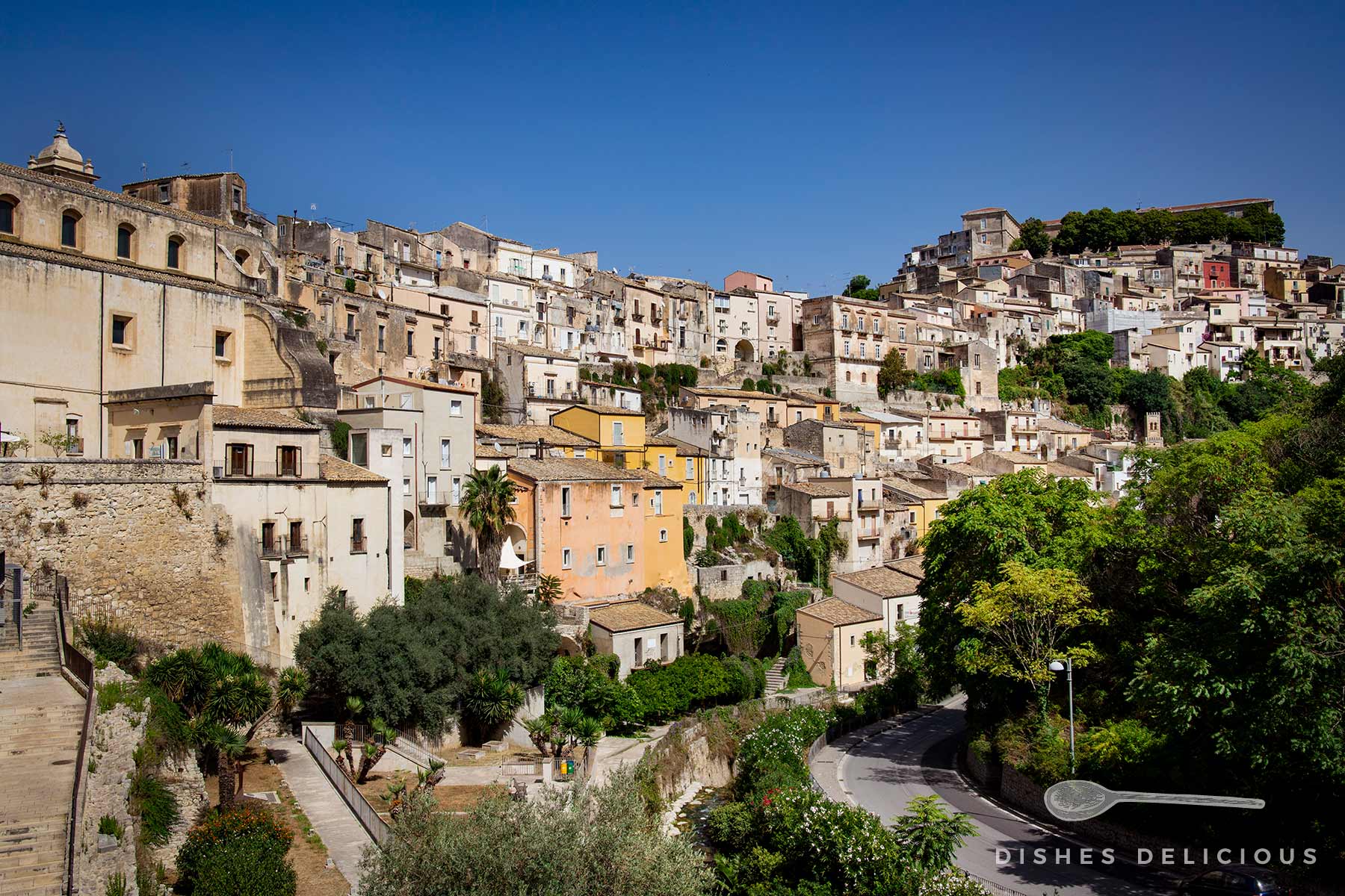 Panorama der dicht aneinander gebaute Häuser in Ragusa Ibla, die den Hang hinaufklettern. Im Vordergrund der Teil einer Steinmauer, eine Treppe und einige Bäume und Sträucher, die ein wenig Grün in die steinerne Umgebung bringen.
