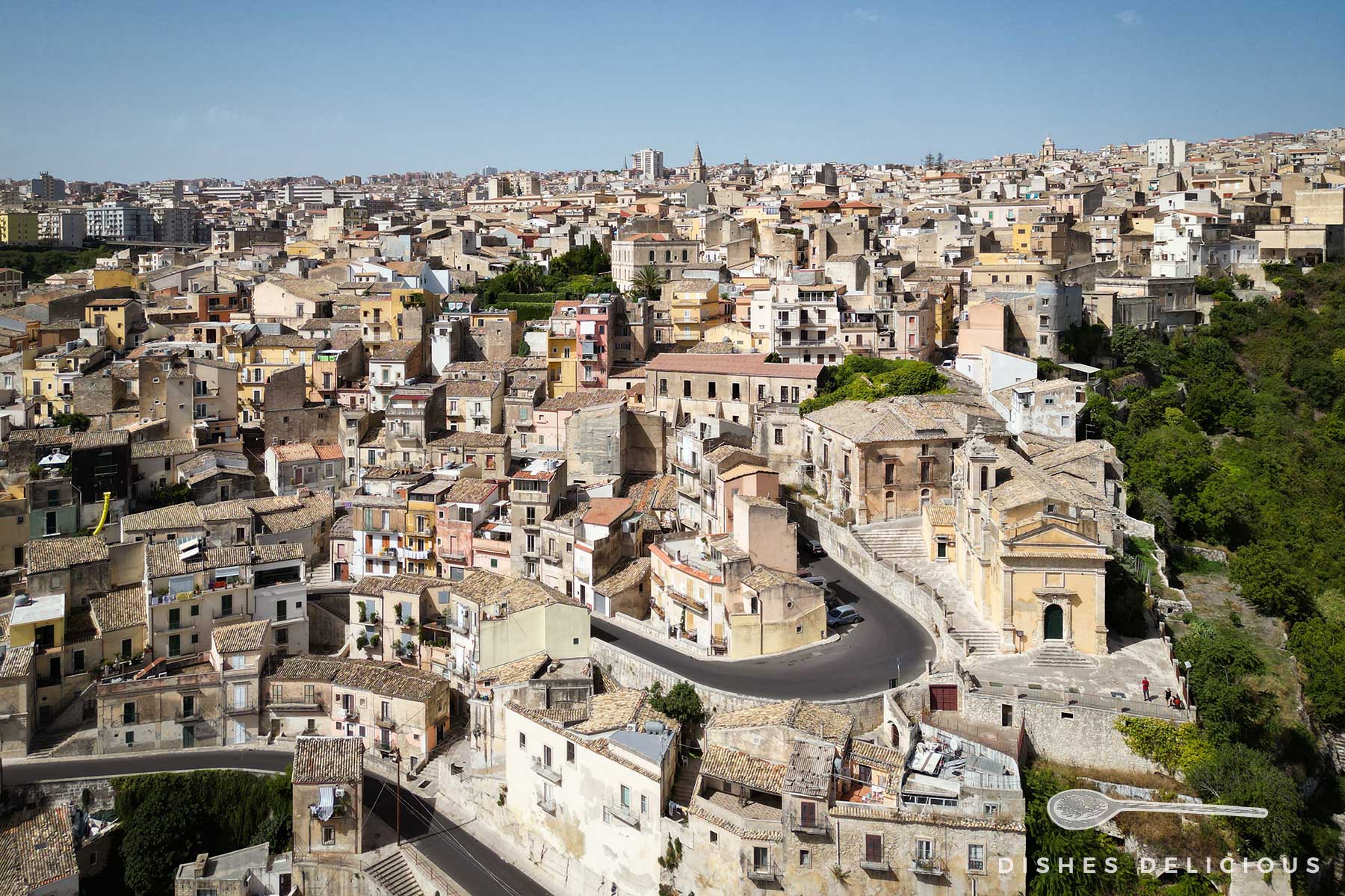 Luftaufnahme von Ragusa Superiore in Sizilien. Häuser erstrecken sich über einen Hügel, den eine kurvige Straße hinaufführt. Rechts im Bild die Kirche Santa Lucia.