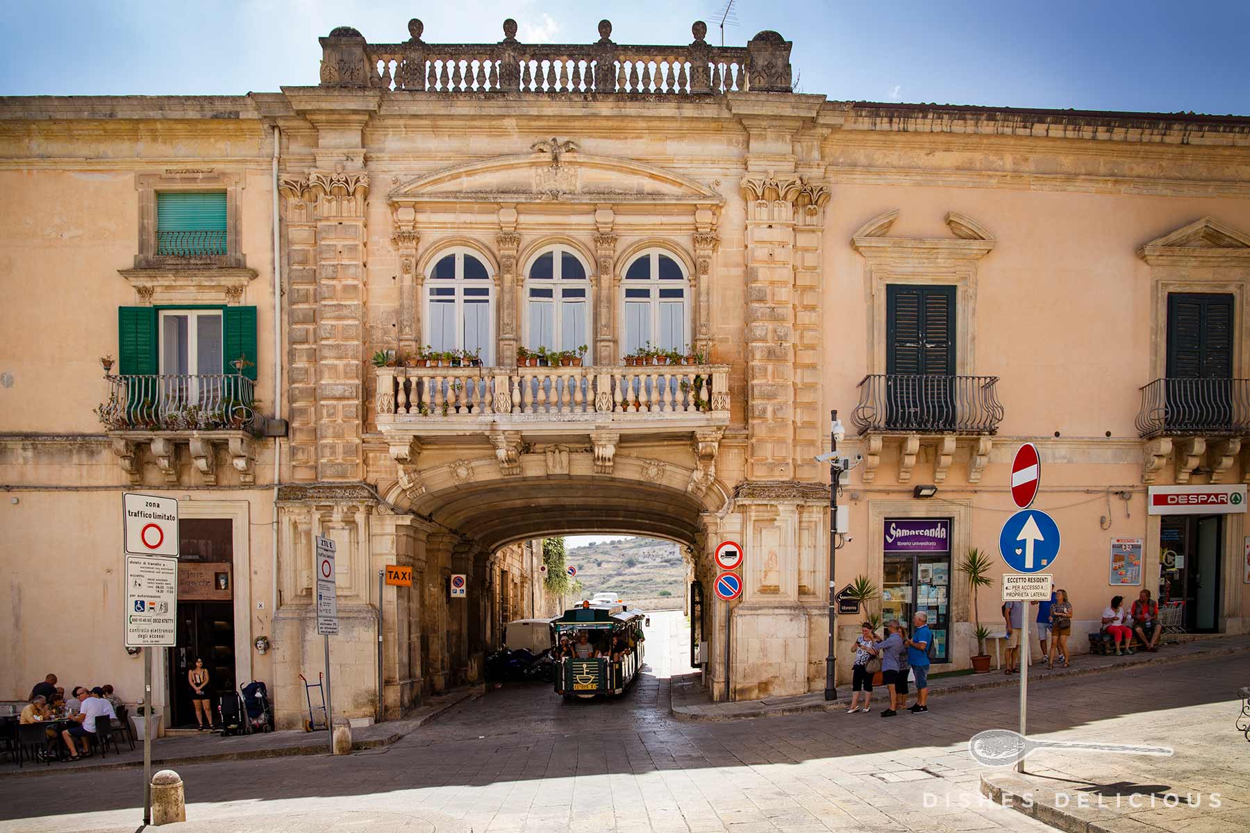 Sehenswürdigkeit Palazzo Arezzo San Filippo in Ragusa Ibla: historisches Barock-Gebäude mit Balkonen und Fenstern. Menschen sind auf der Straße vor dem Gebäude zu sehen. Durch eine Durchfahrt im Gebäude fährt ein Touristen-Bähnchen.