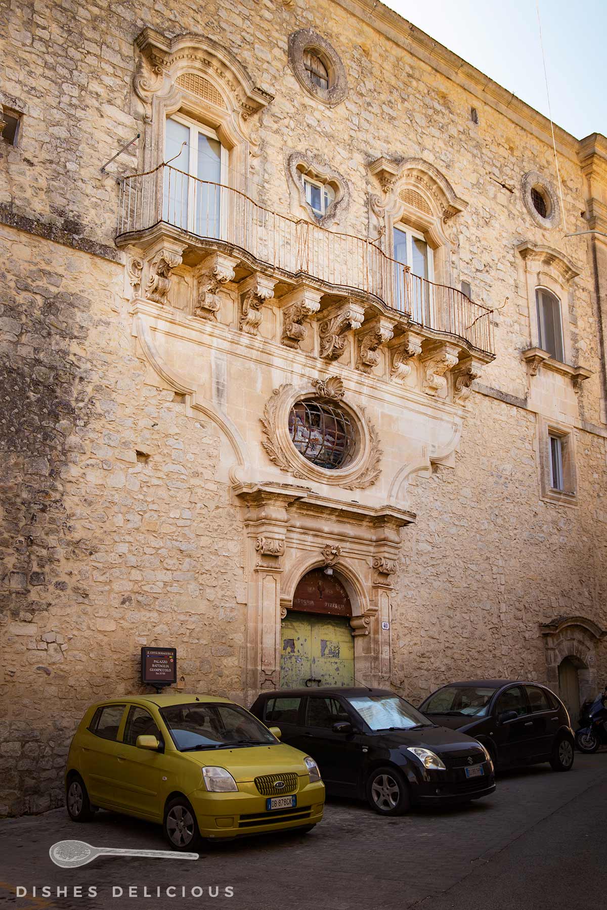 Fassade mit Barockverzierungen des Palazzo Battaglia in Ragusa. Drei Autos parken vor dem Gebäude.