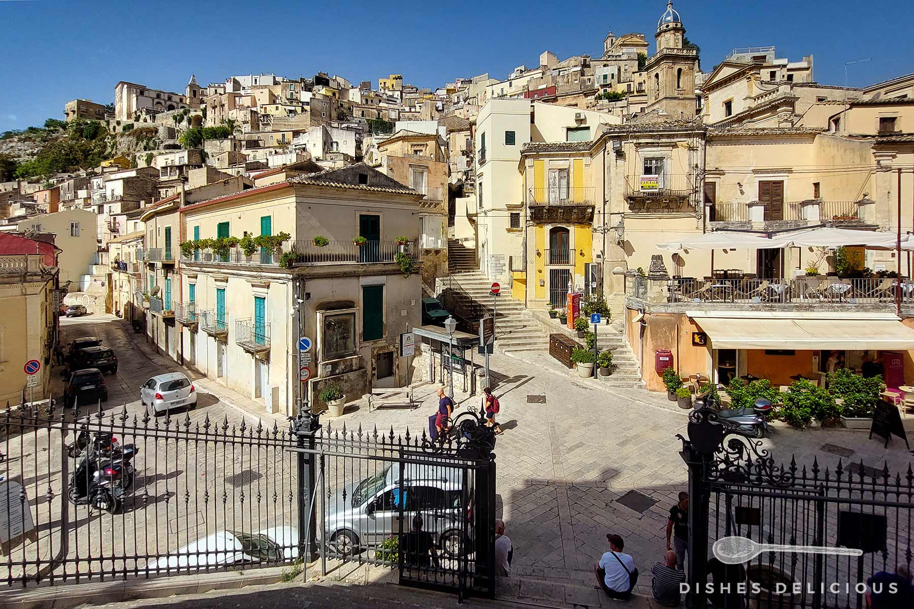 Sonnenbeschienener Piazza della Repubblica in Ragusa, an der sich steile, dicht aneinander gebaute Häuser mit ockerfarbenen, gelben und weißen Fassaden die Hänge hinauf erheben. Ein steiler Treppenaufgang führt von dem Platz zu höher gelegenen Häusern.