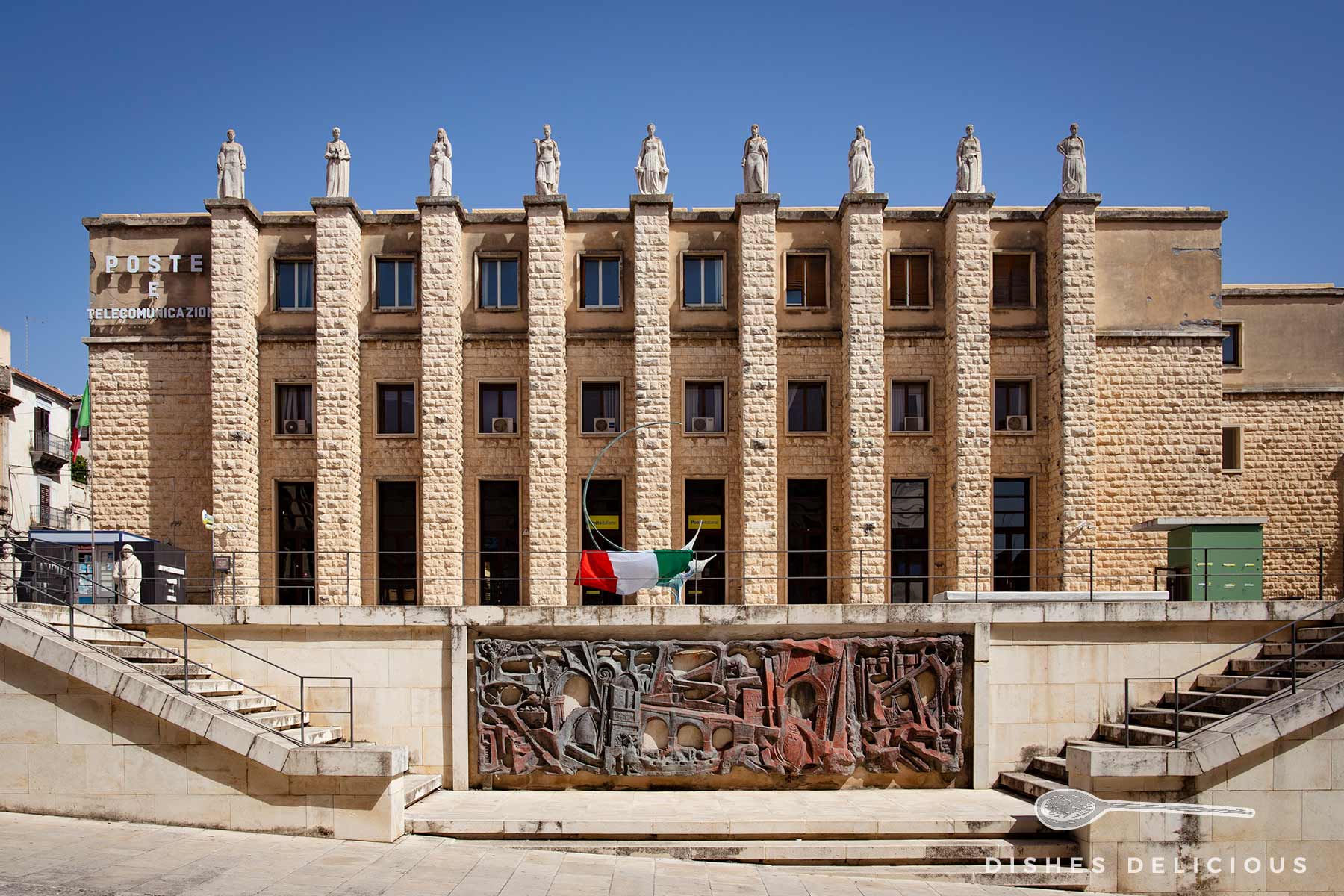 Postpalast von Ragusa - ein typisches Beispiel faschistischer Architektur der 1930er-Jahre mit einer strengen Fassade und Statuen auf dem Dach. Vor dem Gebäude weht die italienische Flagge über einem modernen Relief.
