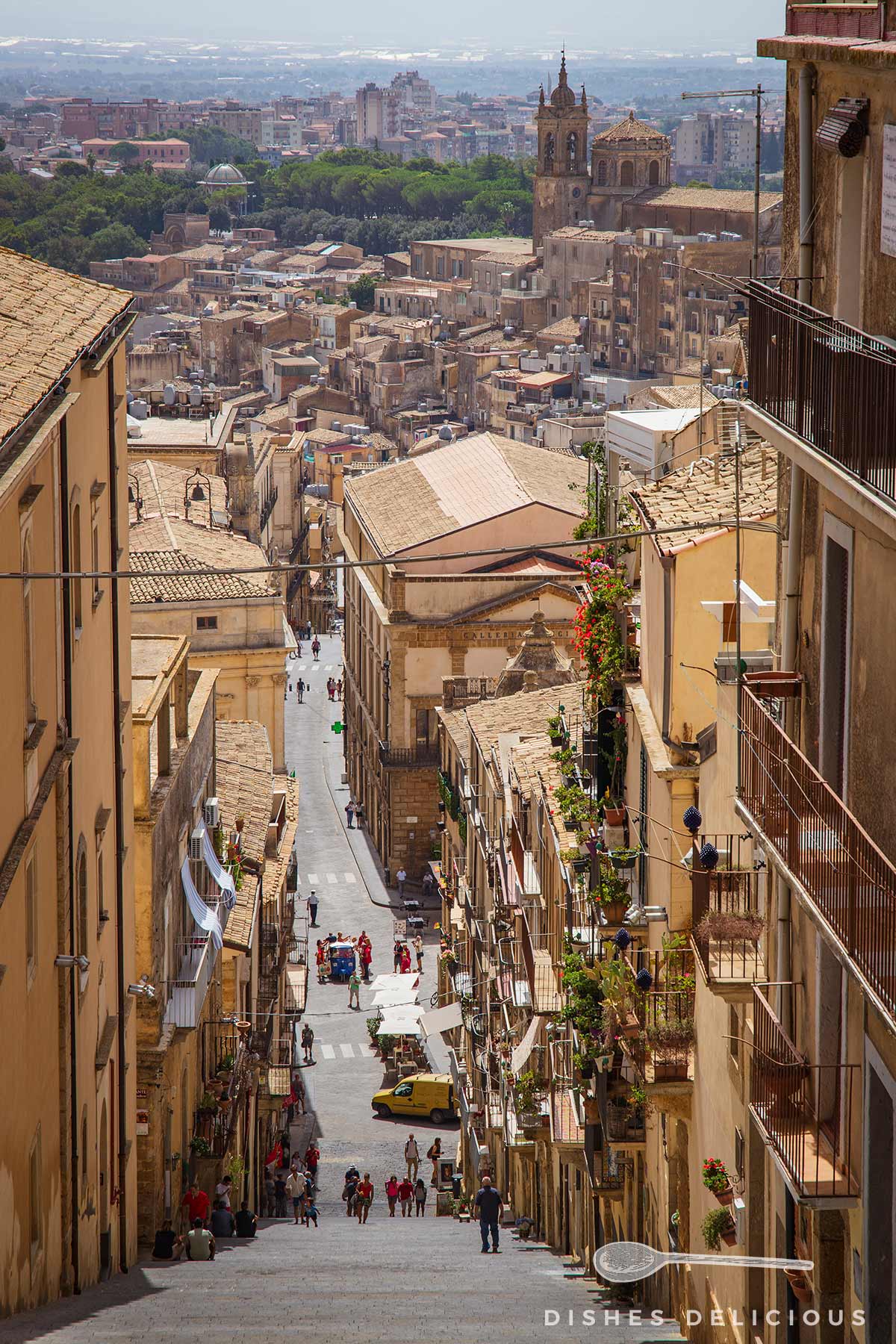 Blick von der Treppe Scalinata Santa Maria del Monte in Caltagirone, Sizilien, auf eine enge Straße mit historischen Gebäuden und einer Kirche im Hintergrund.