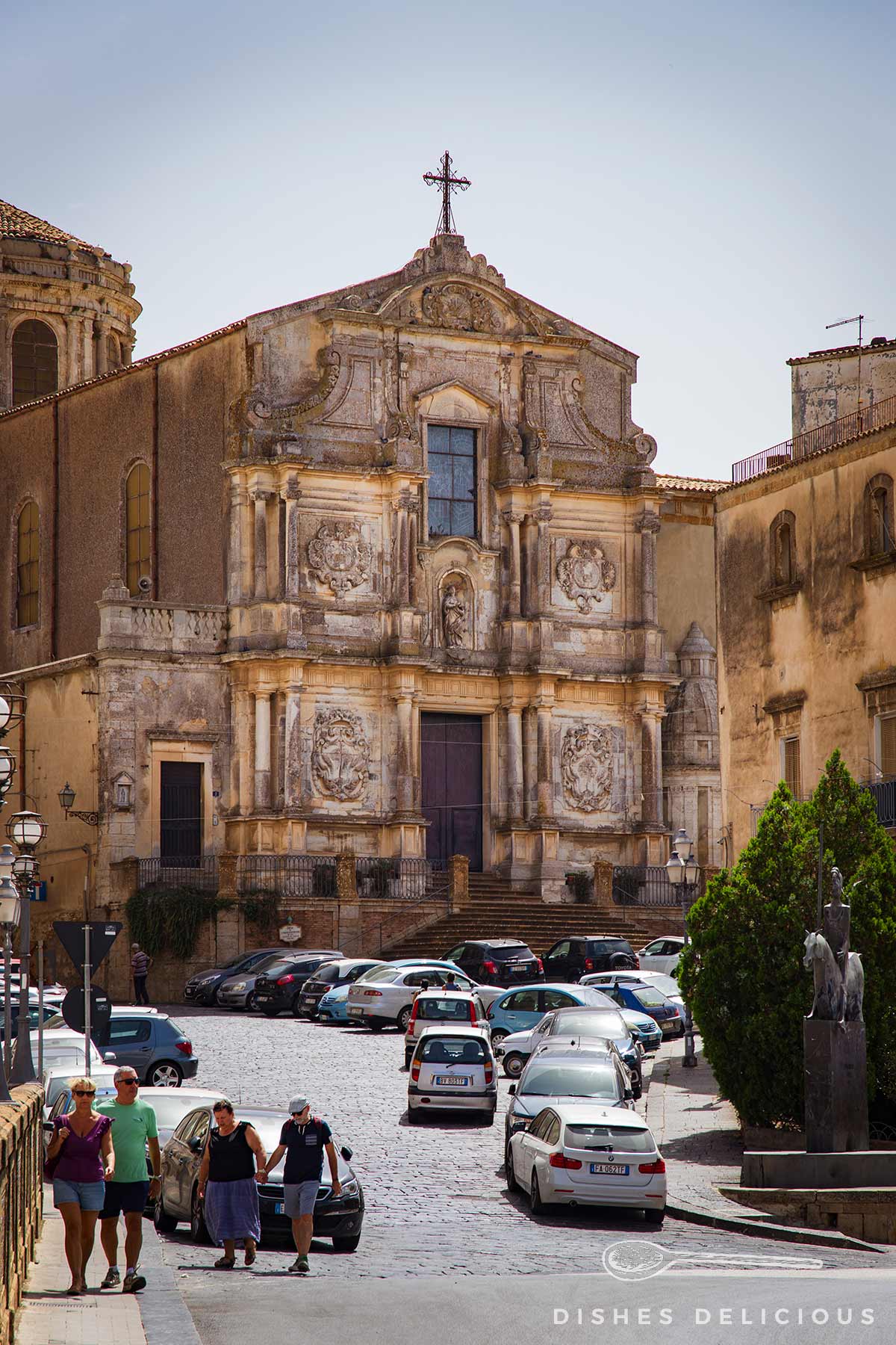 Barocke Fassade der Kirche San Francesco in Caltagirone mit verzierten Reliefs und Kreuz, davor geparkte Autos und Fußgänger auf Kopfsteinpflaster.