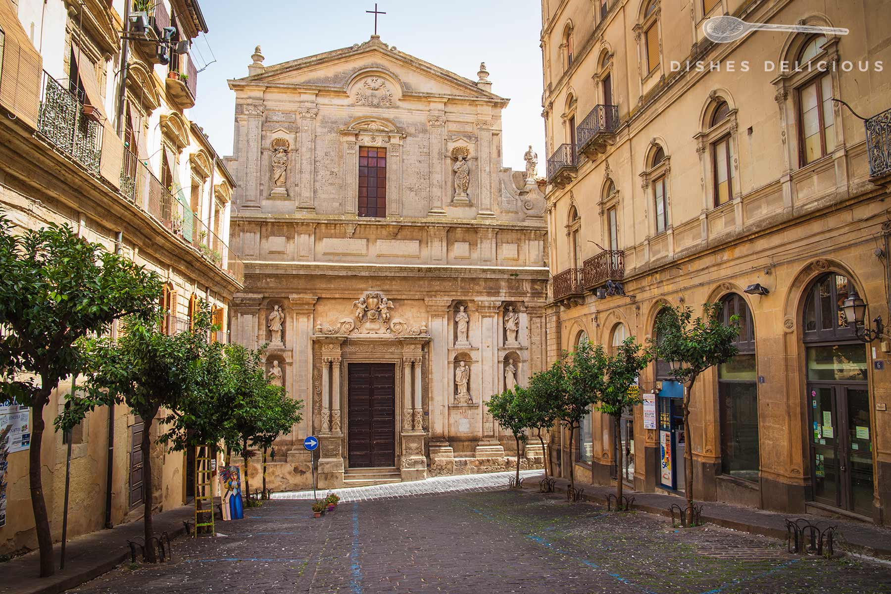 Barocke Fassade der Chiesa del Gesù in Caltagirone mit Statuen und Kreuz, umgeben von historischen Gebäuden und kleinen Bäumen.