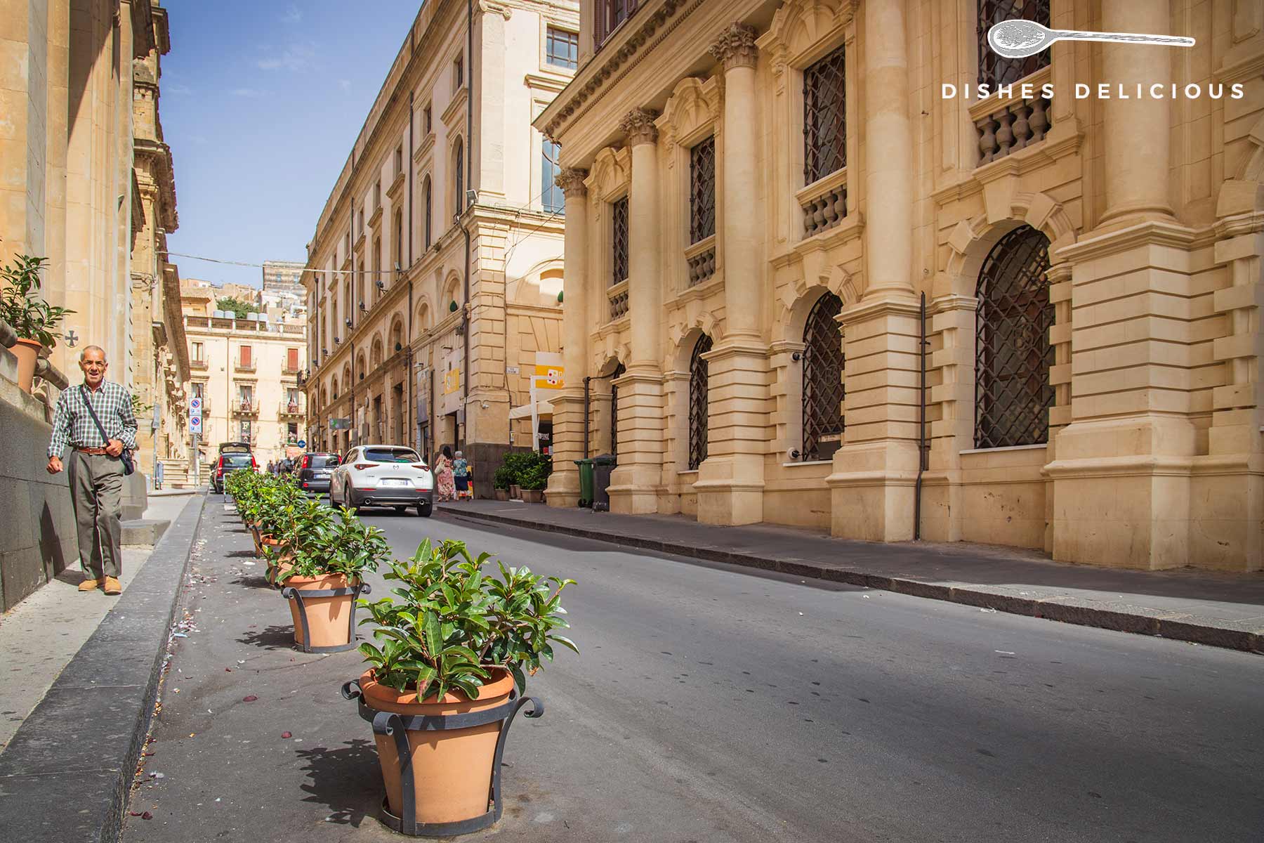 Straße Corso Vittorio Emanuele in Caltagirone mit historischen Gebäuden, Blumentöpfen auf der Straße und einem Mann, der auf dem Bürgersteig geht.