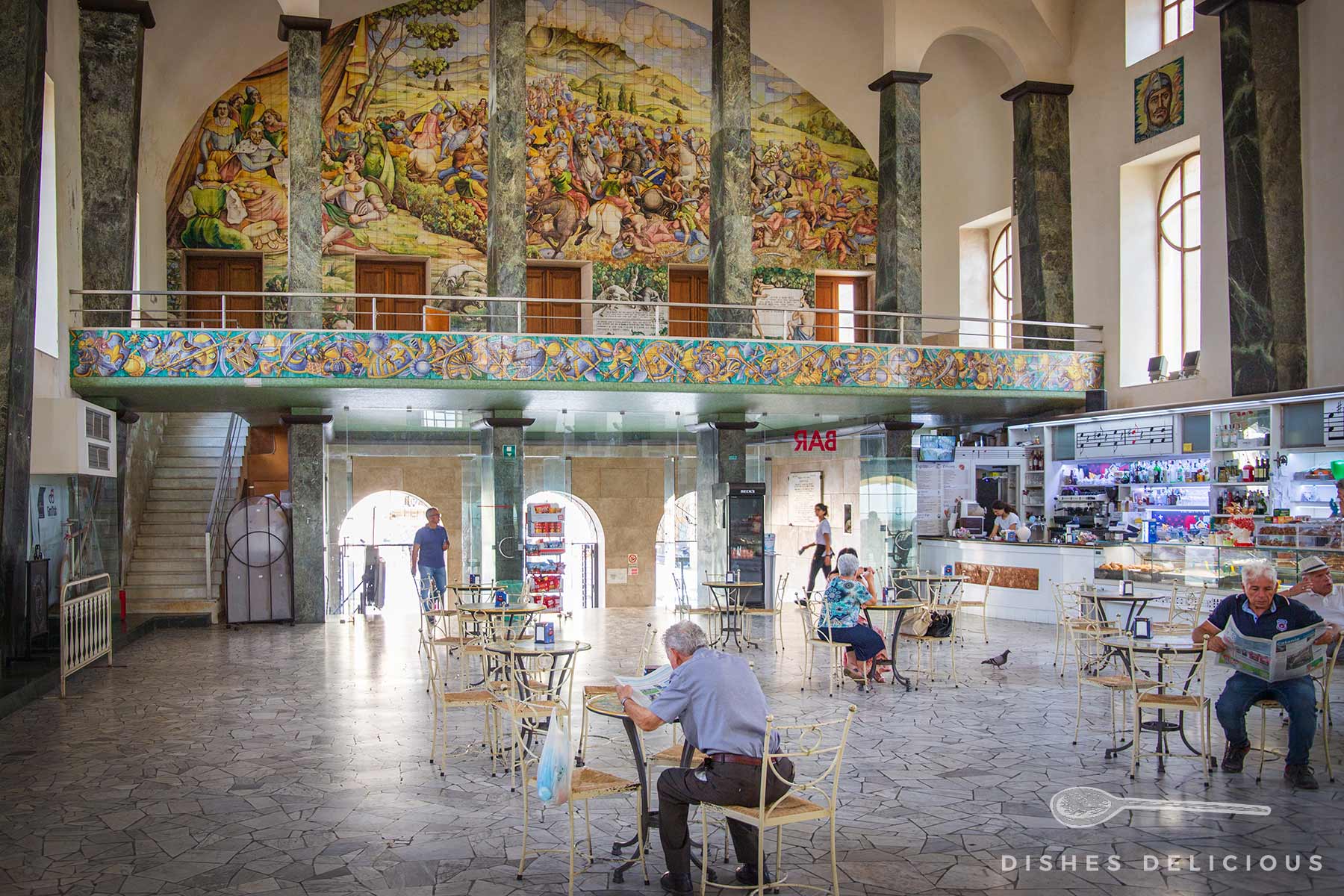 Innenraum der Galleria del Sturzo in Caltagirone mit bunten Wandmalereien, Marmorsäulen, Cafébereich und mehreren älteren Personen an Tischen, die Zeitung lesen.