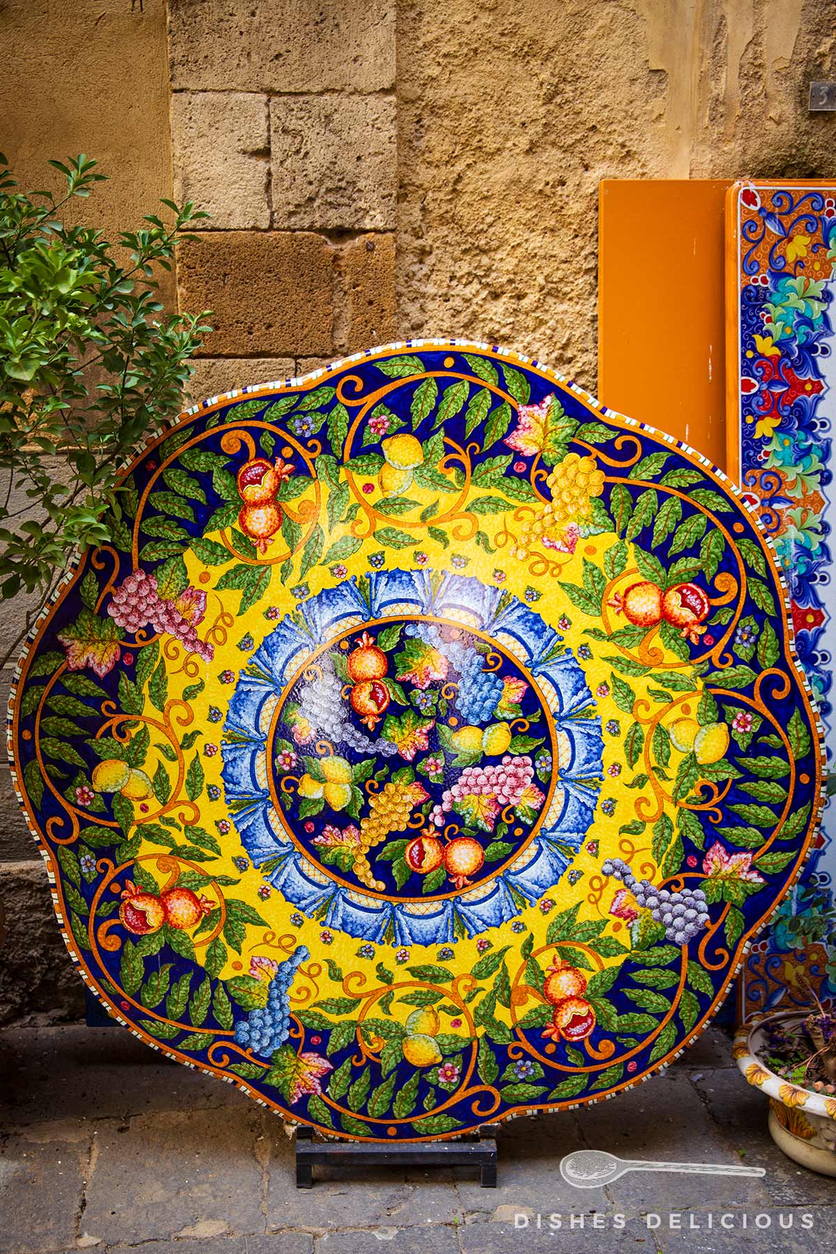 Bunte, runde Keramikplatte mit Frucht- und Blattmotiven, vor einer Steinwand stehend.