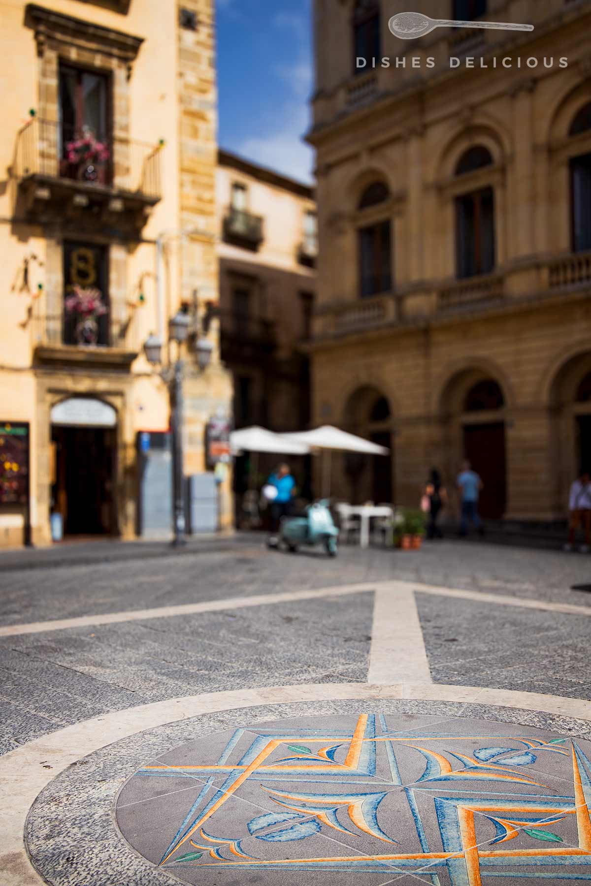 Bunter Mosaikboden mit geometrischem Muster auf dem Piazza Municipio in Caltagirone, umgeben von historischen Gebäuden und Menschen im Hintergrund