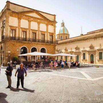 Menschen auf dem sonnigen Piazza del Municipio in Caltagirone mit historischem Gebäude Galleria Luigi Sturzo und der Kathedralen-Kuppel im Hintergrund.