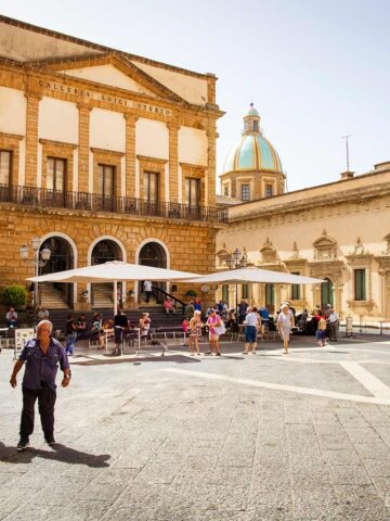 Menschen auf dem sonnigen Piazza del Municipio in Caltagirone mit historischem Gebäude Galleria Luigi Sturzo und der Kathedralen-Kuppel im Hintergrund.