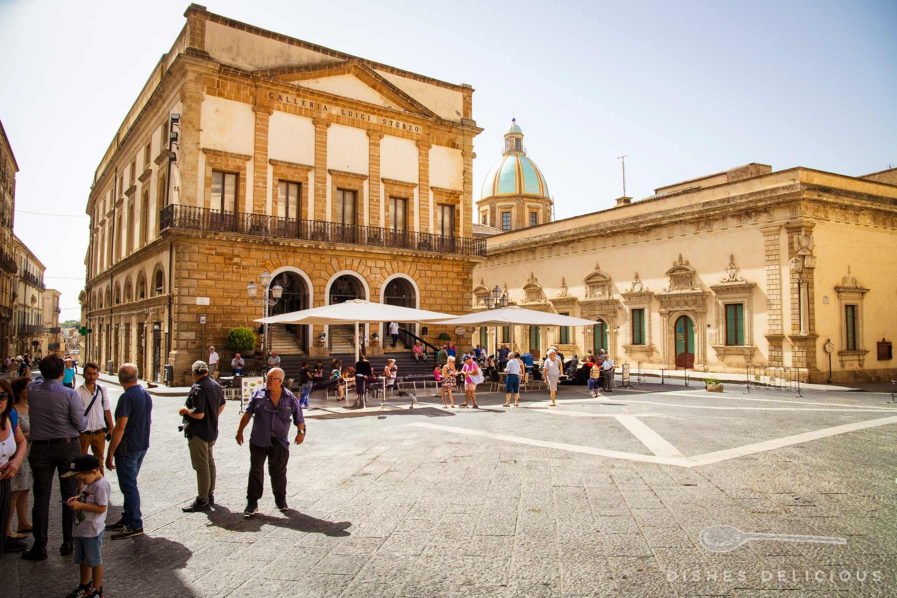 Menschen auf dem sonnigen Piazza del Municipio in Caltagirone mit historischem Gebäude Galleria Luigi Sturzo und der Kathedralen-Kuppel im Hintergrund.