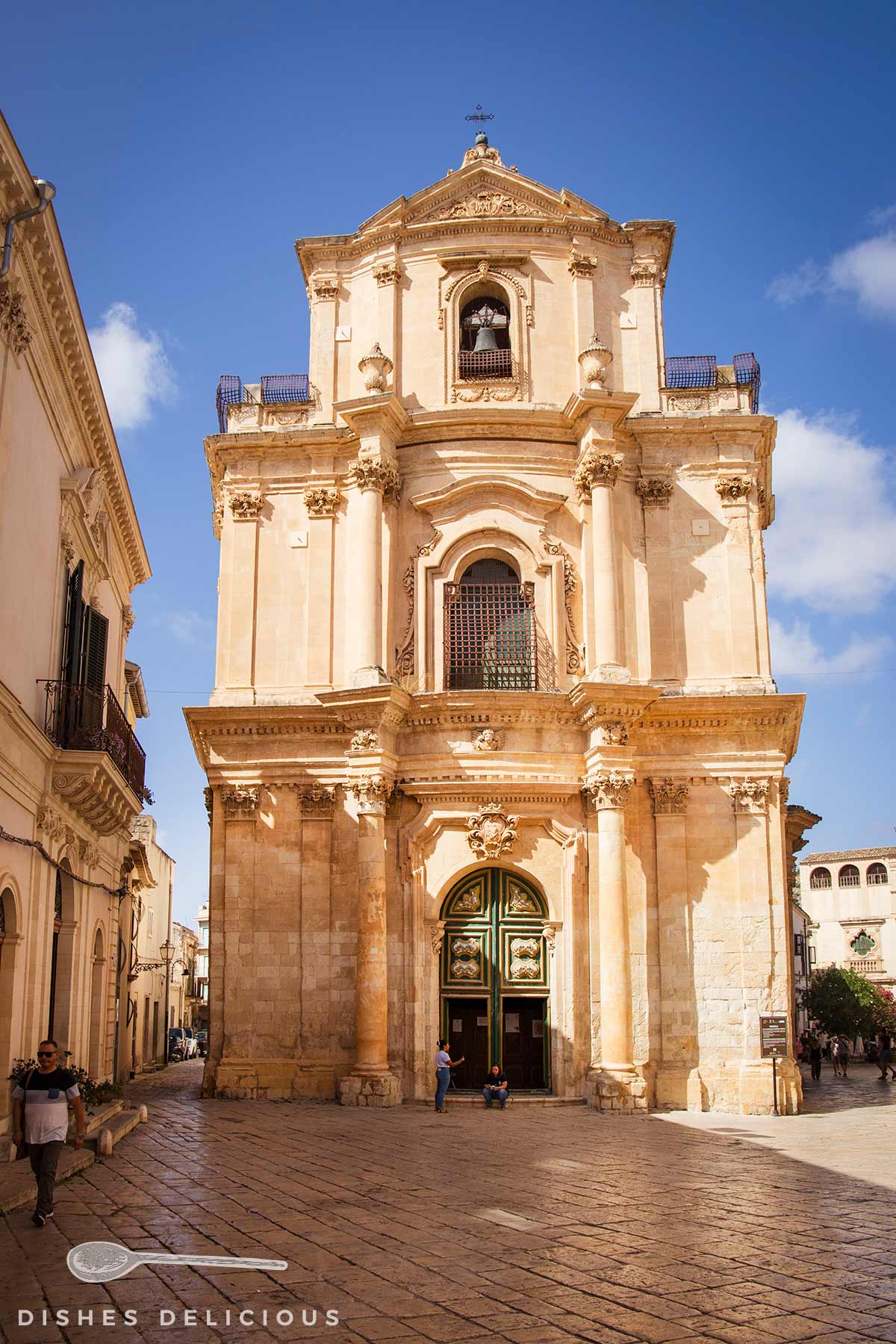 Barocke Fassade der Kirche San Michele Arcangelo in Scicli mit großen Säulen und verzierten Türen unter blauem Himmel.