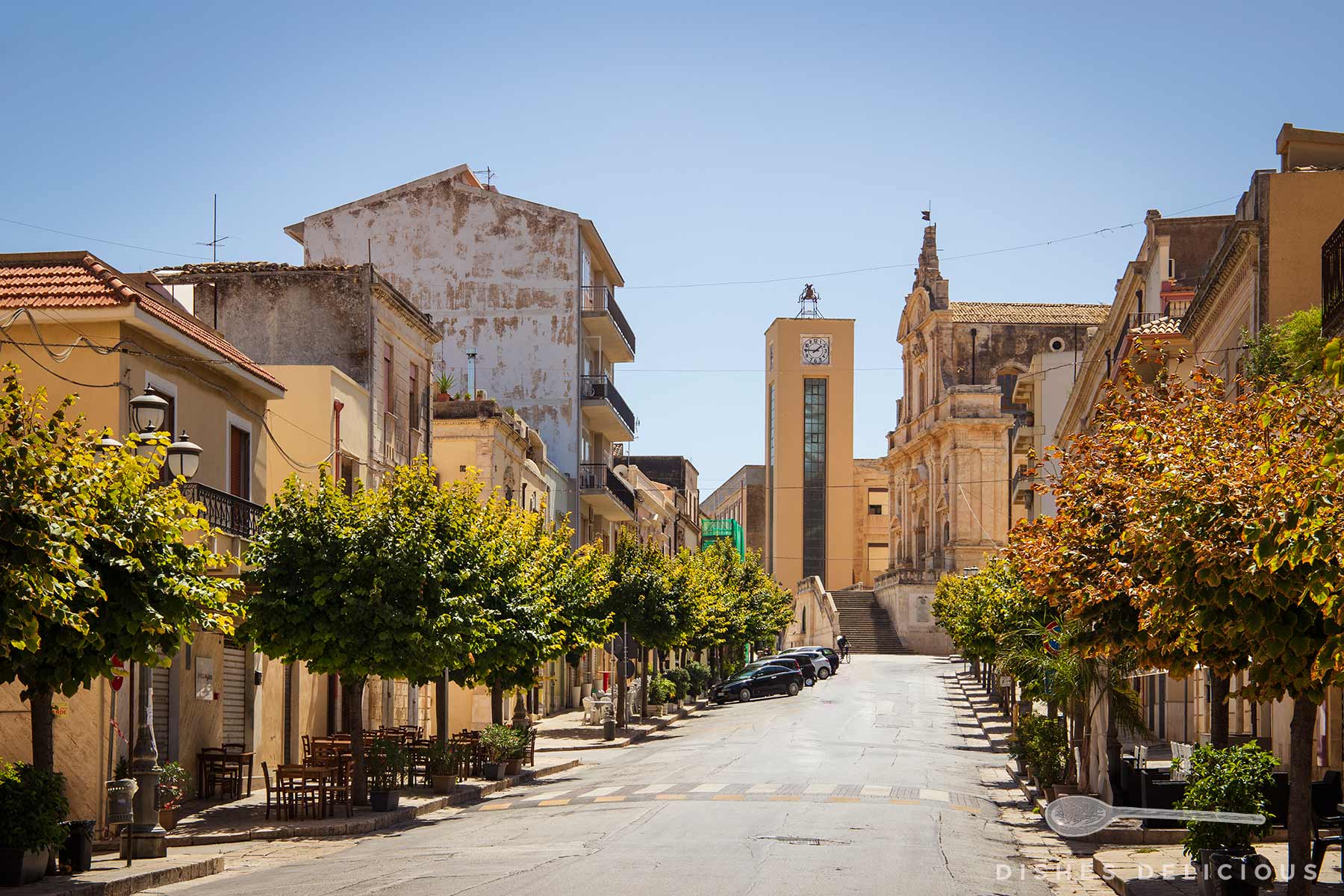 Blick auf die ruhige Straße Corso Garibaldi in Ispica mit Bäumen, Parkplätzen und historischen Gebäuden, darunter eine Kirche und ein Uhrturm im Hintergrund.
