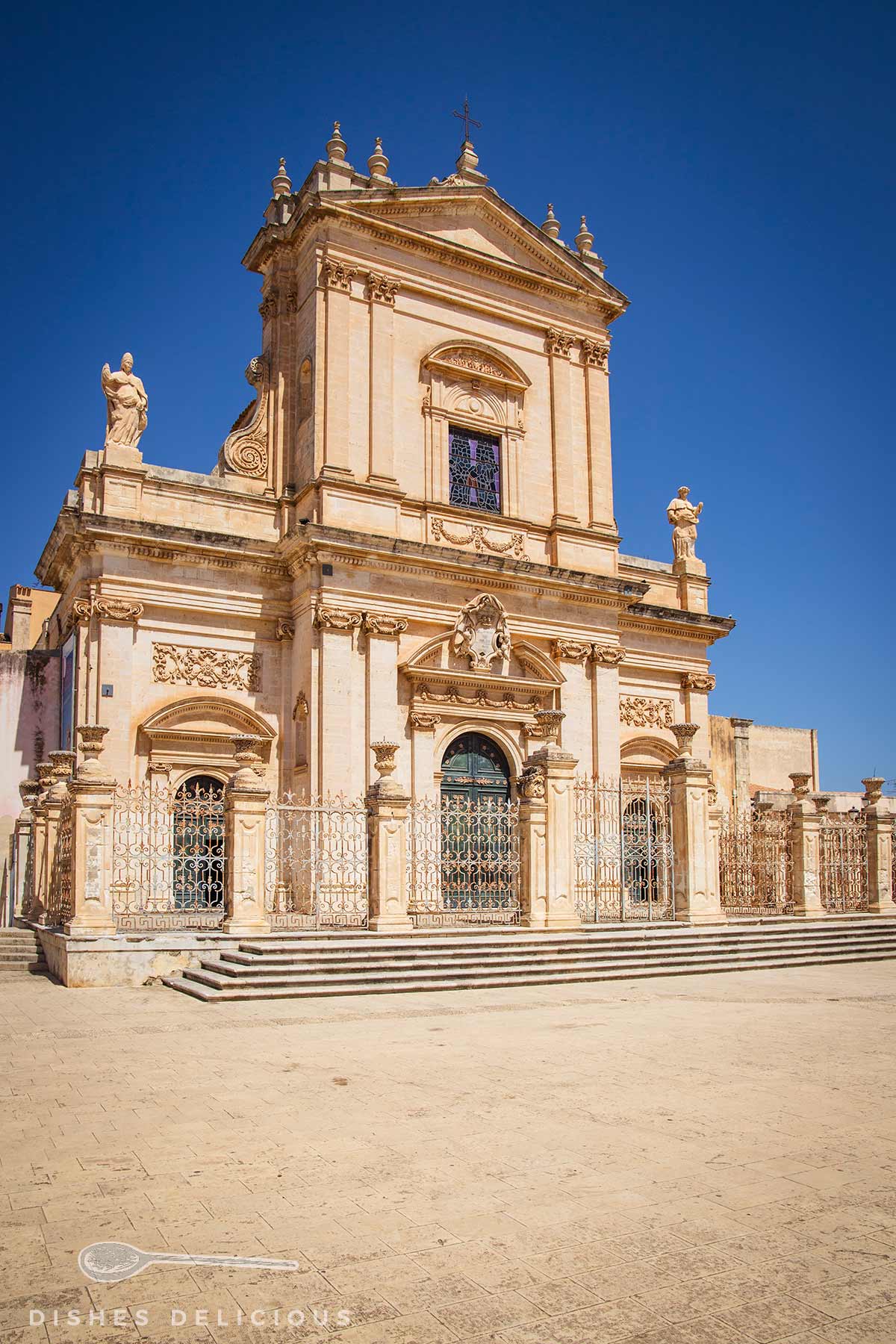 Fassade der Basilica Santa Maria Maggiore in Ispica mit barocker Architektur, Statuen und einem großen Eingangstor unter klarem blauem Himmel