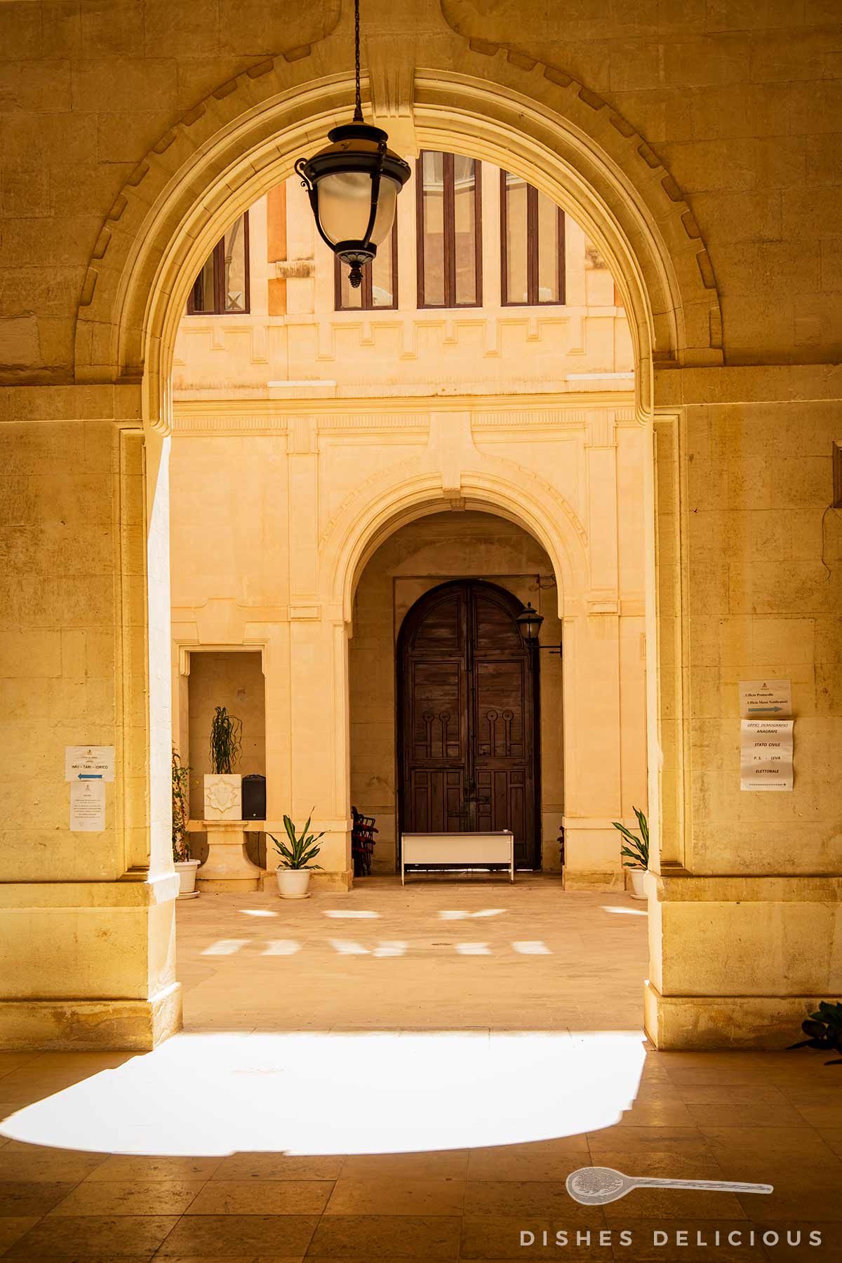 Innenhof des Palazzo Belmonte mit hellen Steinbögen, hängender Laterne und dunkler Holztür im Hintergrund.