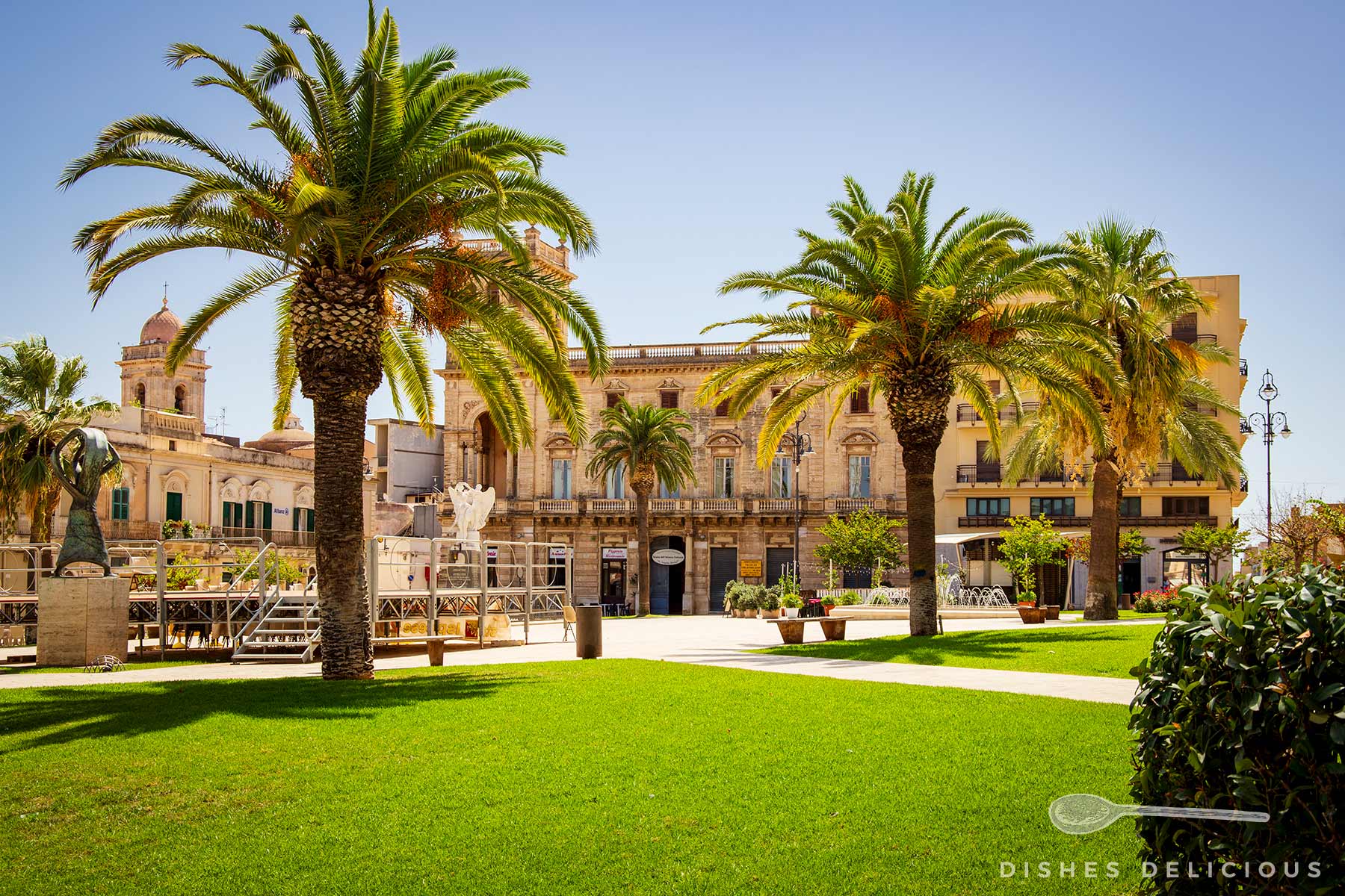 Piazza l'Unità Italia in Ispica mit grüner Rasenfläche, Palmen, historischen Gebäuden und Skulpturen bei strahlend blauem Himmel.