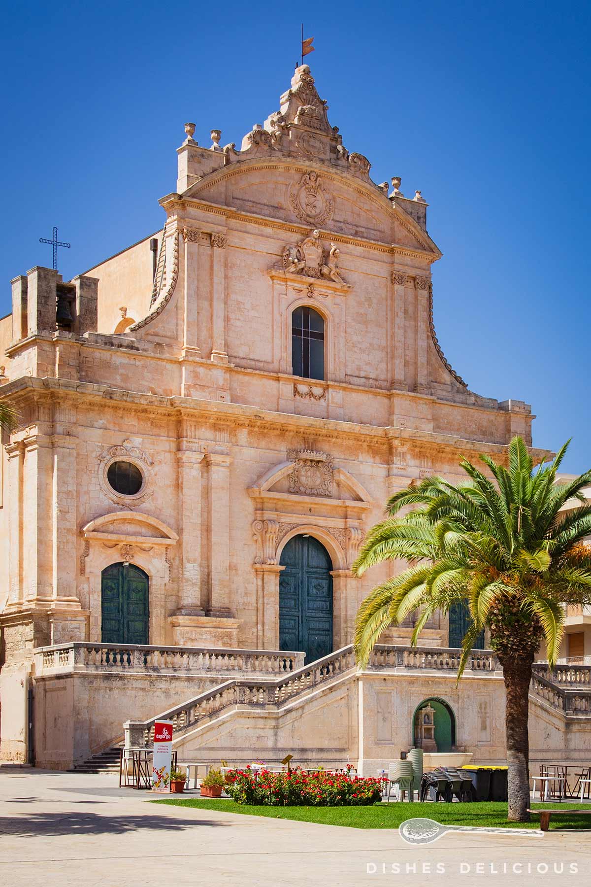 Barockkirche San Bartolomeo in Ispica mit aufwendiger Fassade, großer Treppe und Palme im Vordergrund.