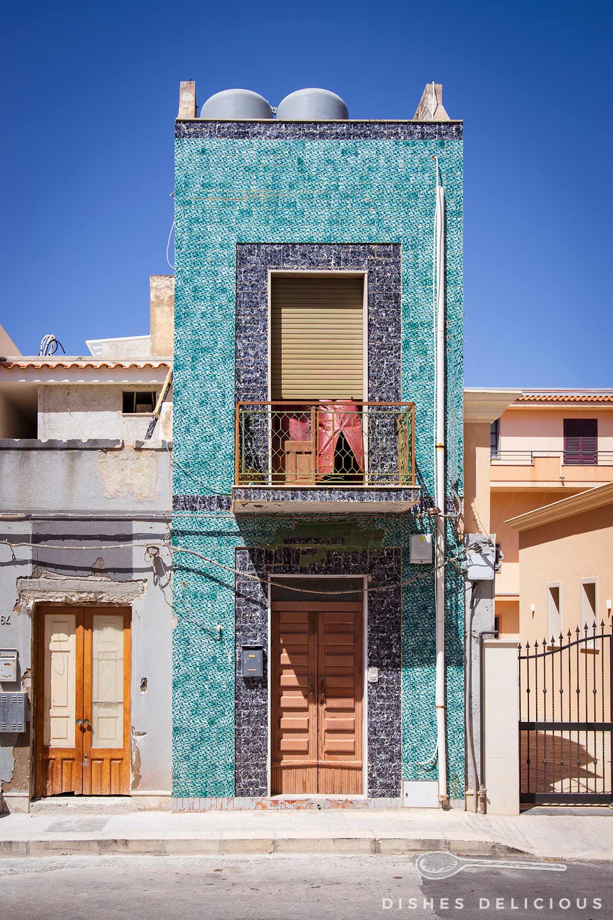 Zweistöckiges Wohnhaus in Ispica mit türkisfarbenen Mosaikfliesen und braunen Holztüren unter klarem blauem Himmel.