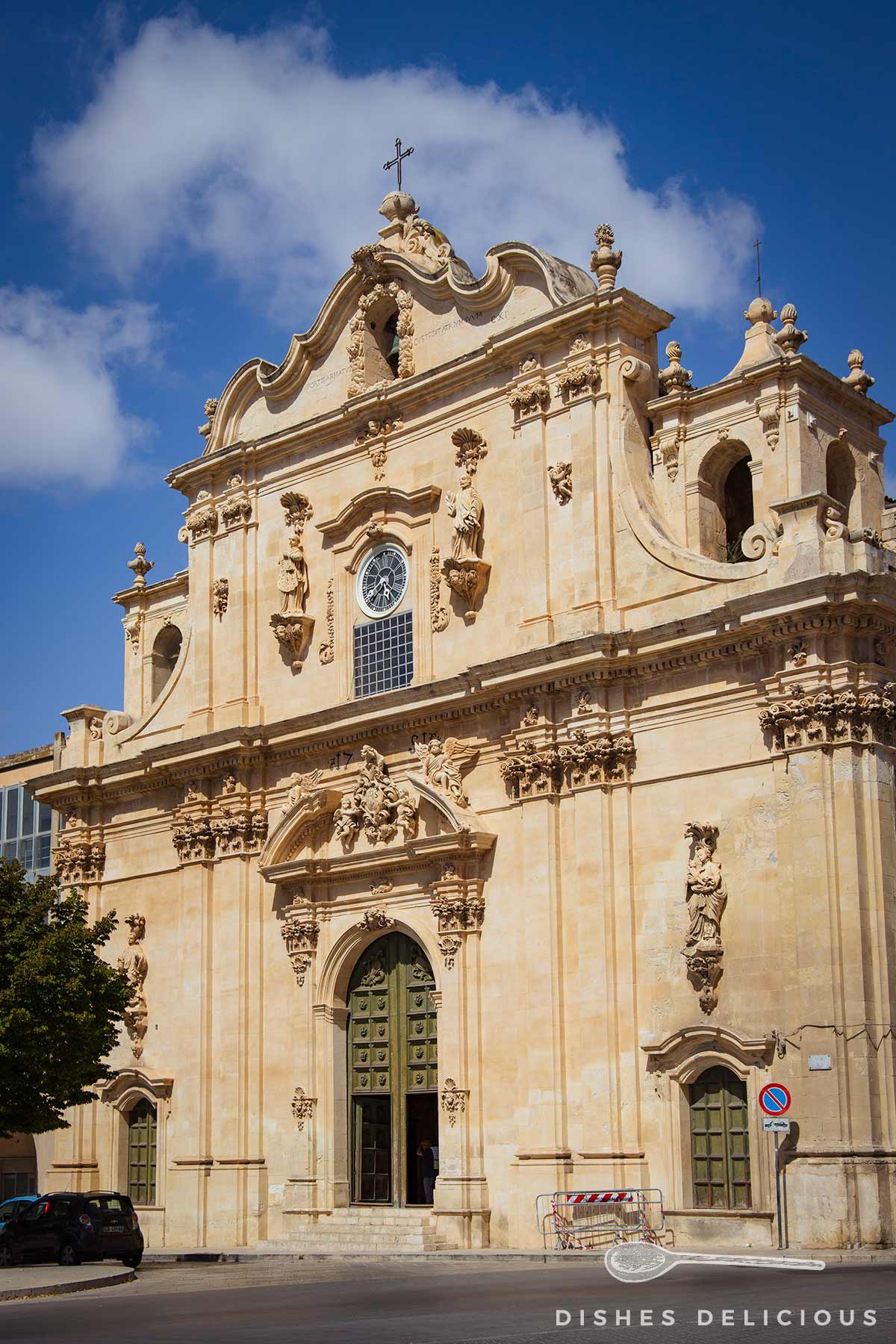 Barocke Fassade der Mutterkirche von Scicli, die mit Statuen geschmückt ist. Die Türen am großen Eingangsportal sind geöffnet.
