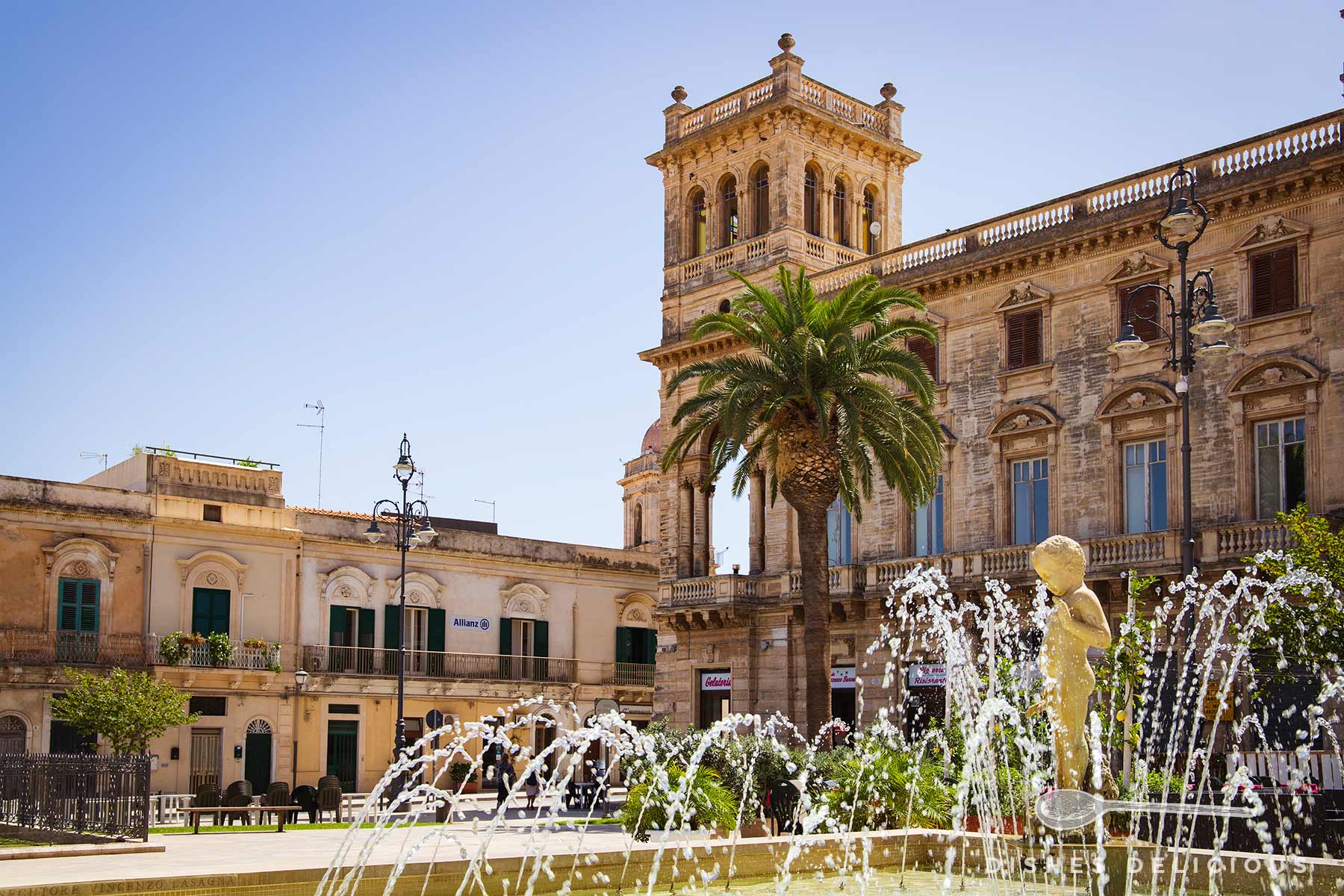 Palazzo Bruno mit einem Brunnen im Vordergrund, der eine Statue eines Kindes zeigt, umgeben von Wasserstrahlen und einer Palme vor dem historischen Gebäude.