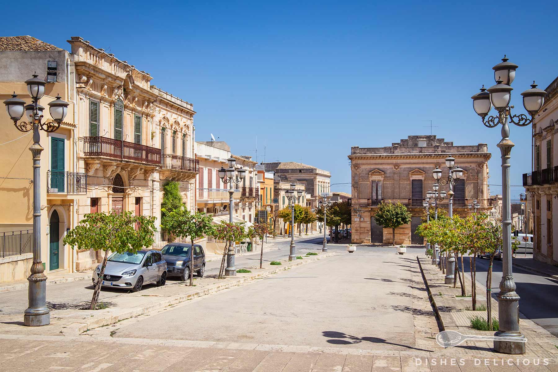 Piazza Annunziata in Ispica mit historischen Palazzi gesäumt, kleinen Bäumen und Straßenlaternen unter klarem blauem Himmel.