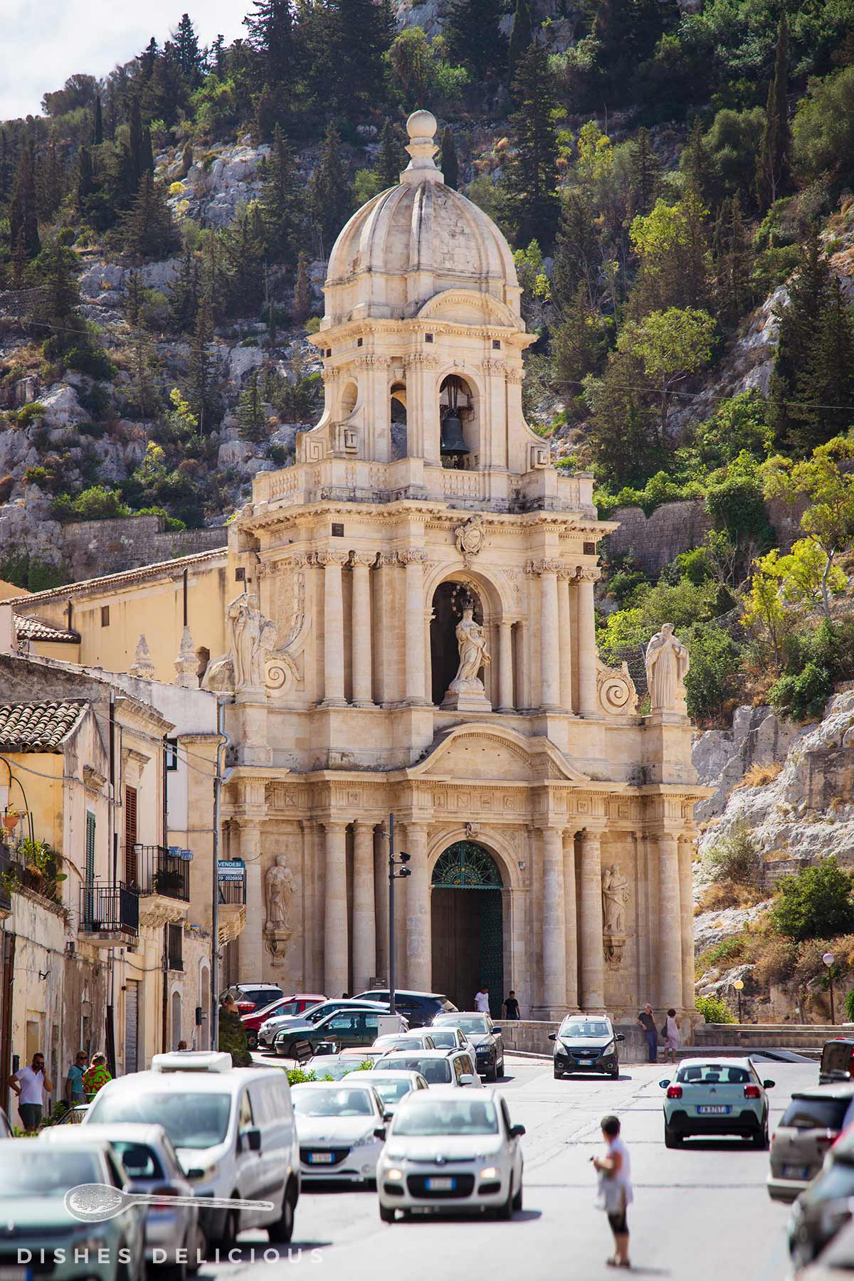 Barocke Fassade der Kirche San Bartolomeo in Scicli mit Glockenturm und Statuen, umgeben von bewaldeten Hügeln und belebter Straße mit Autos.