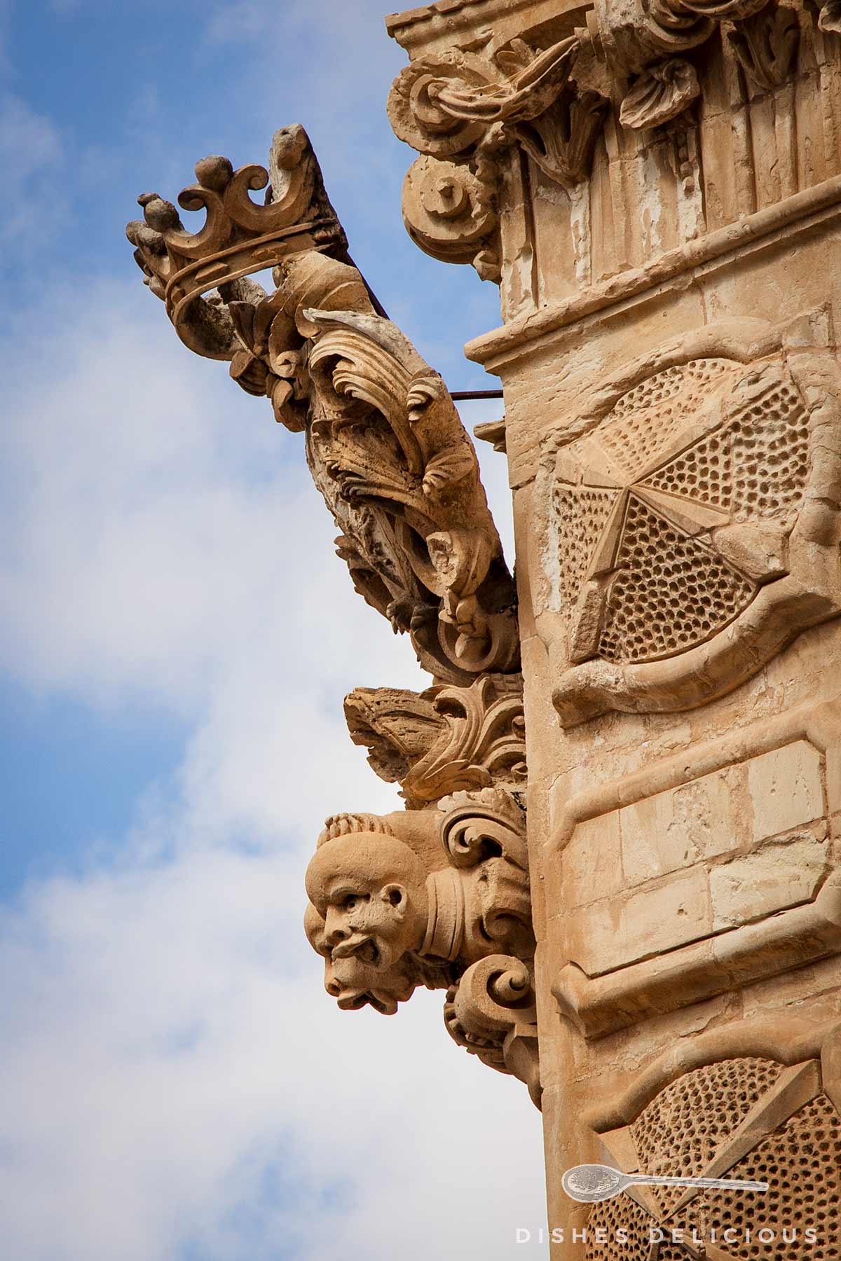 Detailreiche Steinverzierungen an der Fassade des Palazzo Beneventano in Scicli mit einem Kopf und einer Krone vor blauem Himmel