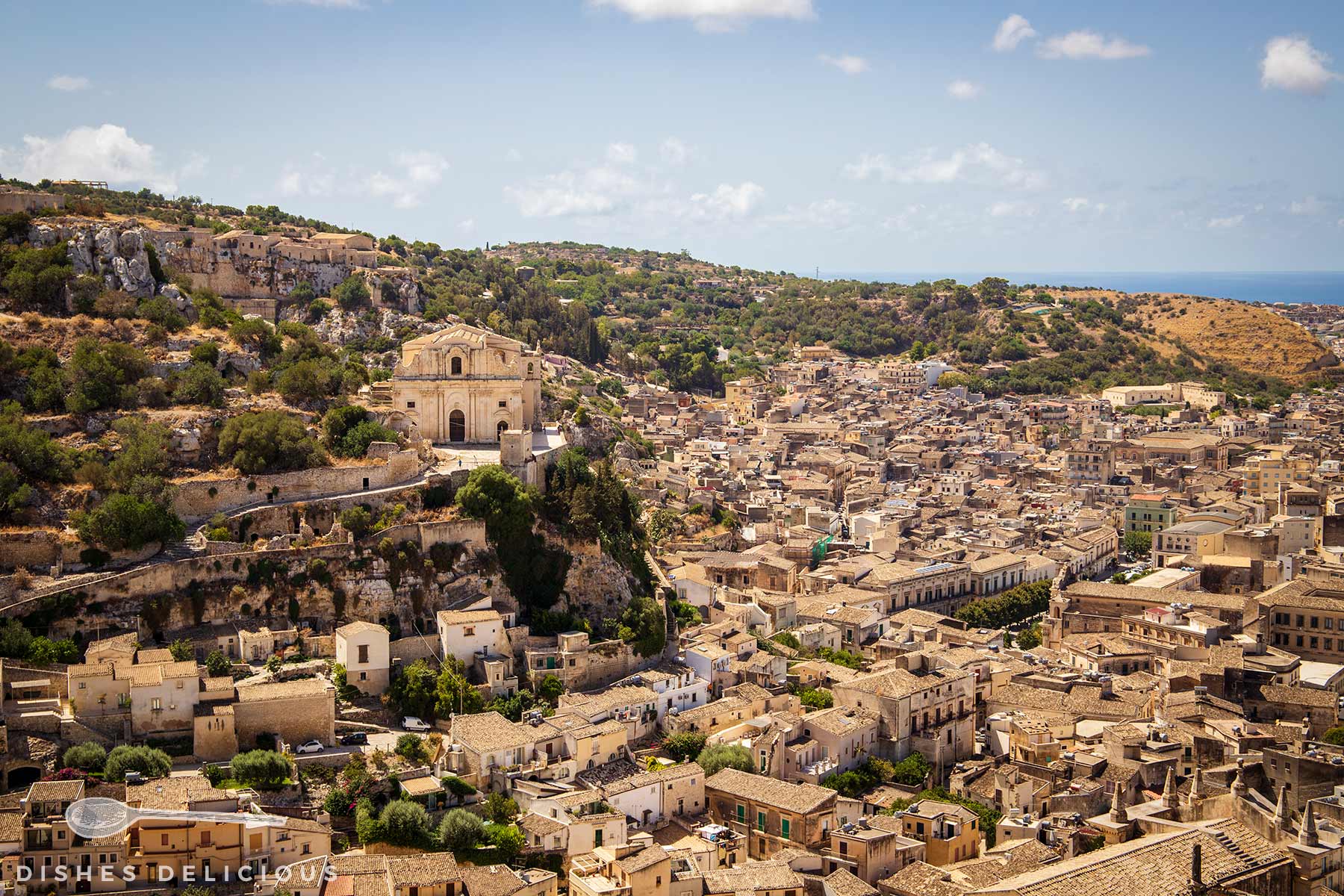 Blick auf die Stadt Scicli in Sizilien mit barocker Kirche auf einem Hügel und dicht bebauten Häusern unter blauem Himmel, das Meer im Hintergrund.