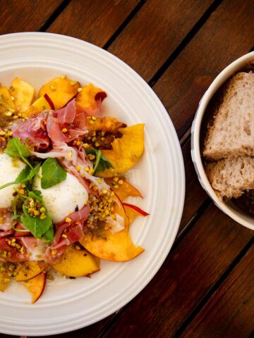 Ein großer Teller mit Pfirsich-Carpaccio mit Schinken und Burrata, garniert mit Rucola und gehackten Pistazien. Daneben eine kleine Schüssel mit geschnittenem Brot.