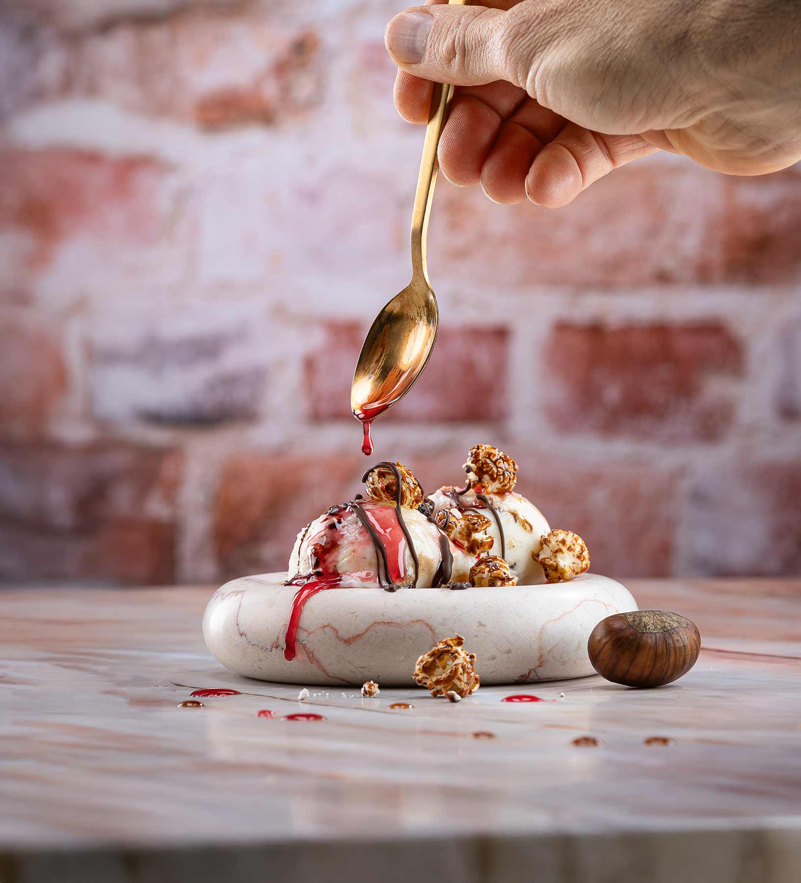 Jemand träufelt mit einem Löffeln Amarena-Kirschsauce auf Vanilleeis mit Maronen und Schoko-Popcorn auf weißem Teller